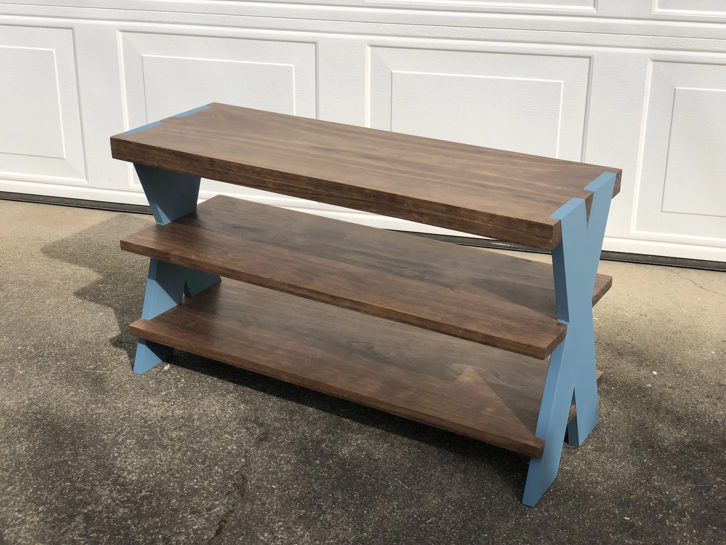 A three-tier wooden shelf unit with blue supports, placed outdoors against a white paneled wall.
