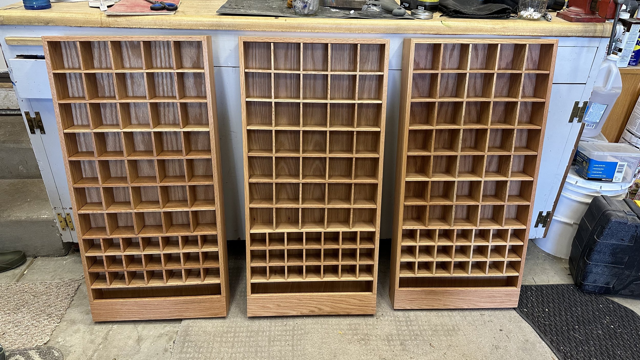 Three wooden shelving racks with multiple small square compartments stand side by side in a workshop or garage setting.