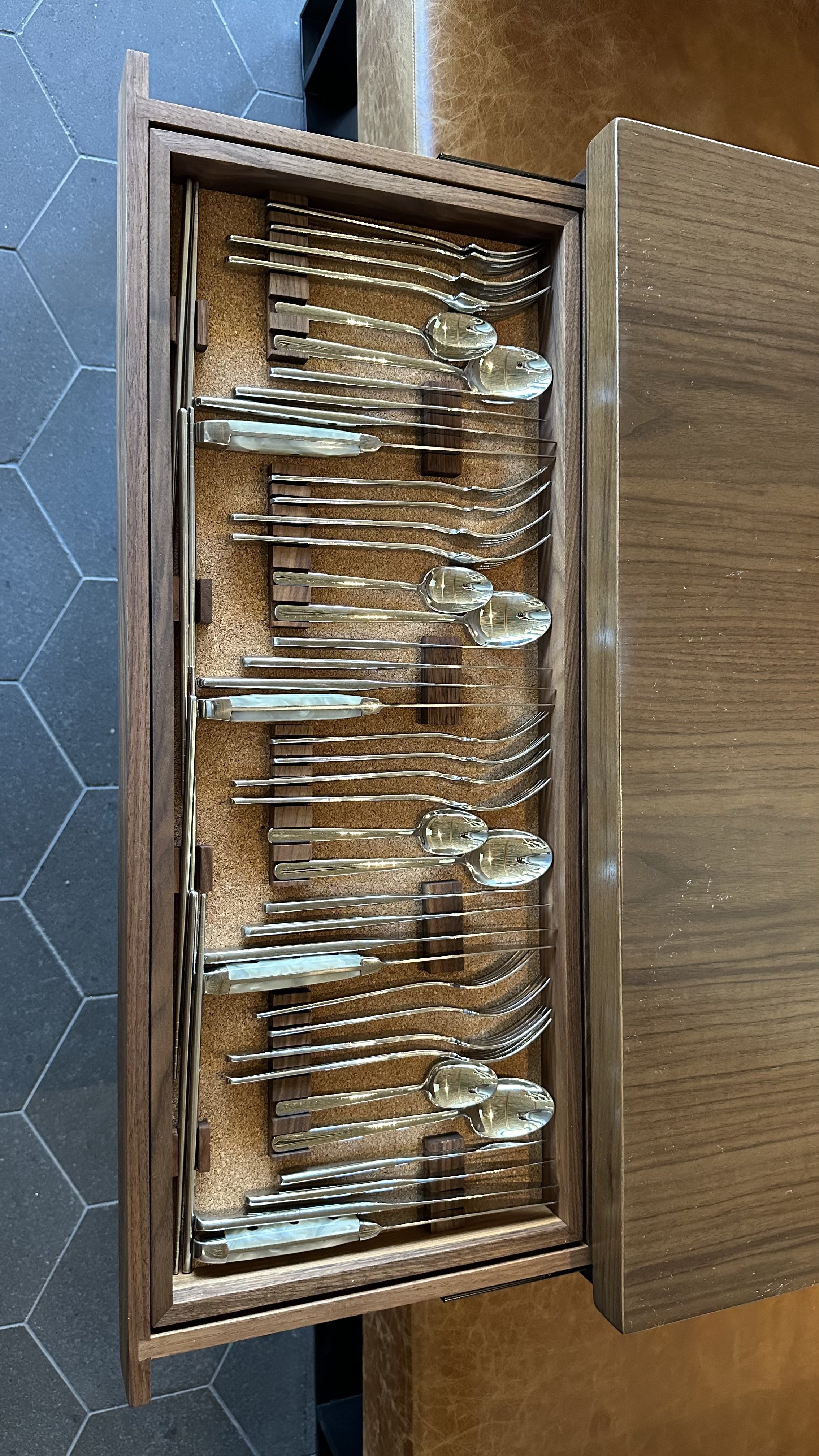 A wooden display case holding a collection of silverware, including forks, knives, and spoons, with a cork lining inside the case. The case is open, revealing the organized silverware inside.