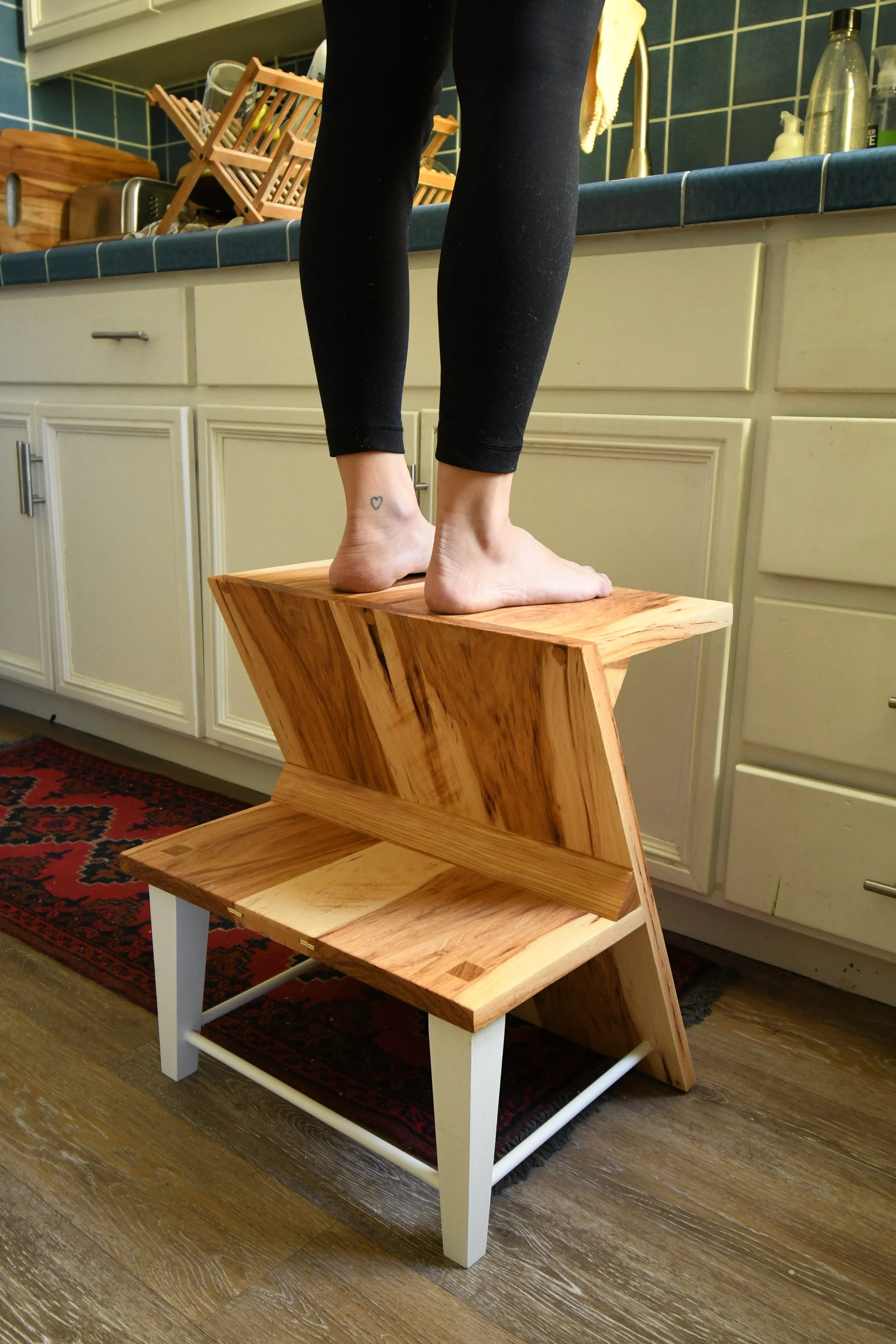 A person standing on a wooden step stool in a kitchen, with their legs and feet visible and a tattoo of a heart on one ankle. The kitchen has white cabinets, a blue tiled countertop, and various kitchen items.