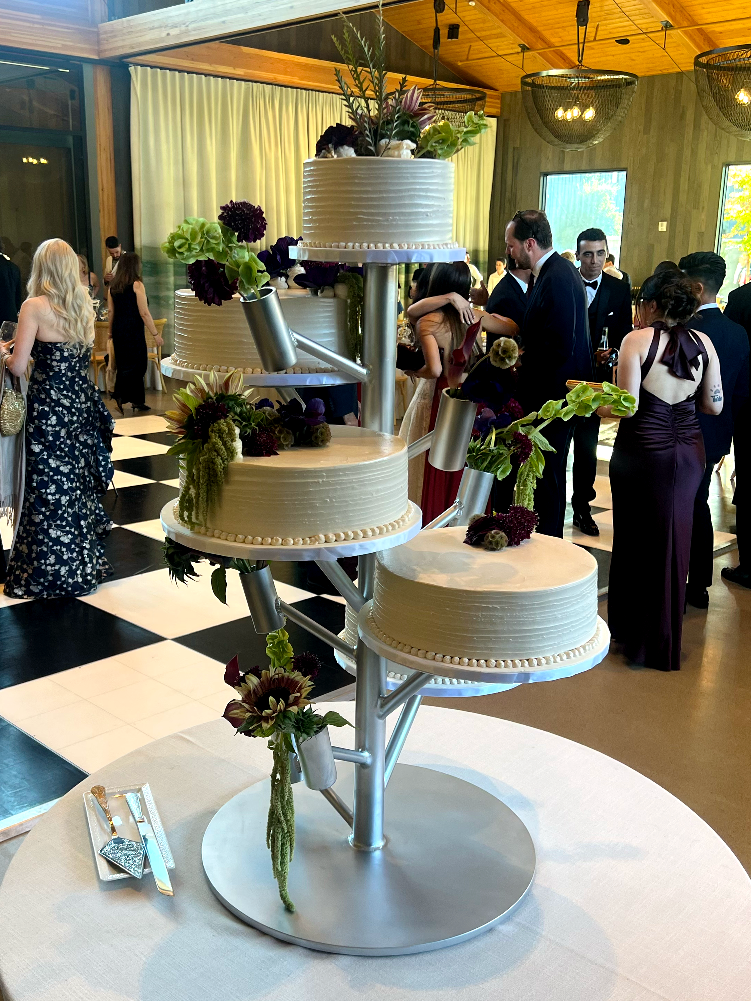 A tiered wedding cake with white frosting, adorned with flowers and greenery, displayed on a table at wedding reception.