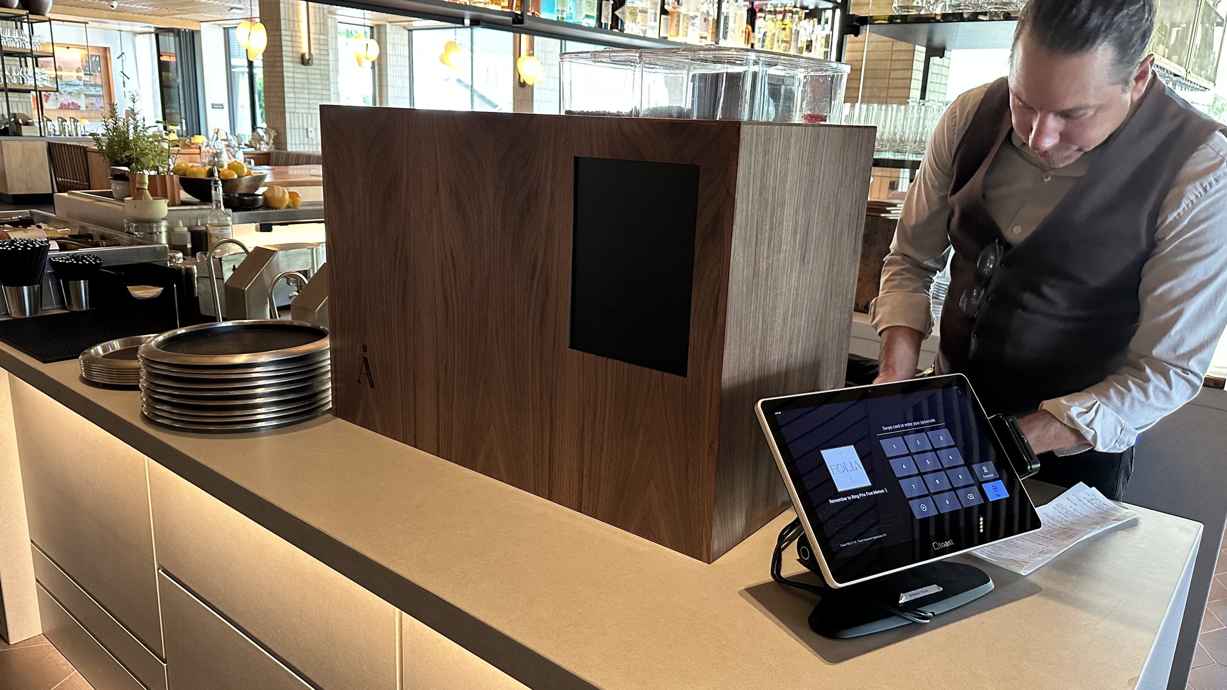 Inside a modern restaurant or cafe, a man is working at a counter with a wooden divider in the foreground. The counter has stacks of plates, a tablet, and a point-of-sale device. In the background, there are dining tables, colorful hanging lights, an
