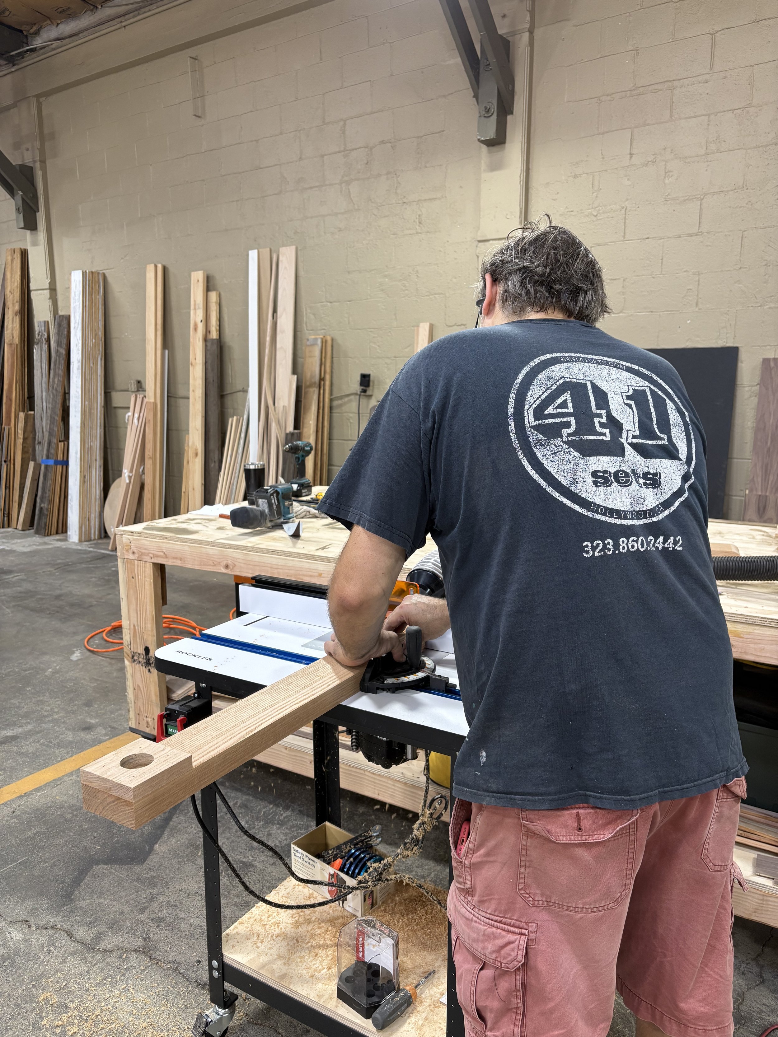 A person working with a woodworking tool on a wooden piece in a workshop, with various wood pieces and tools in the background.