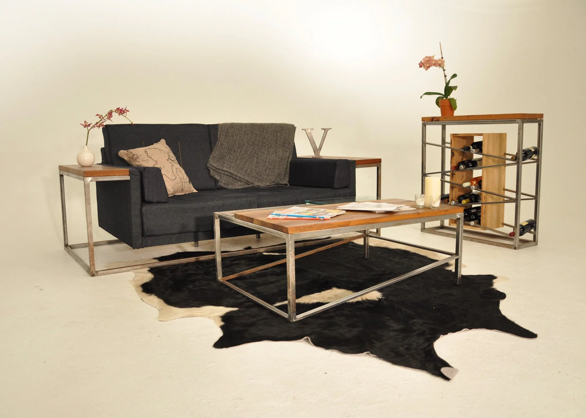 A modern living room setup with a black sofa, a wooden conference table on a black cowhide rug, a matching wooden side table with a white vase and pink orchid, and a wine rack with bottles and a small lamp, all against a plain light-colored wall.