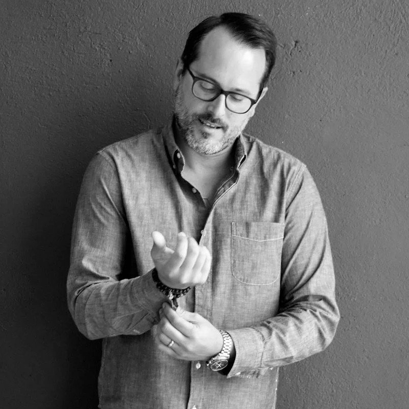 A man with glasses and a beard wearing a button-up shirt, looking down and smiling, standing against a dark wall.