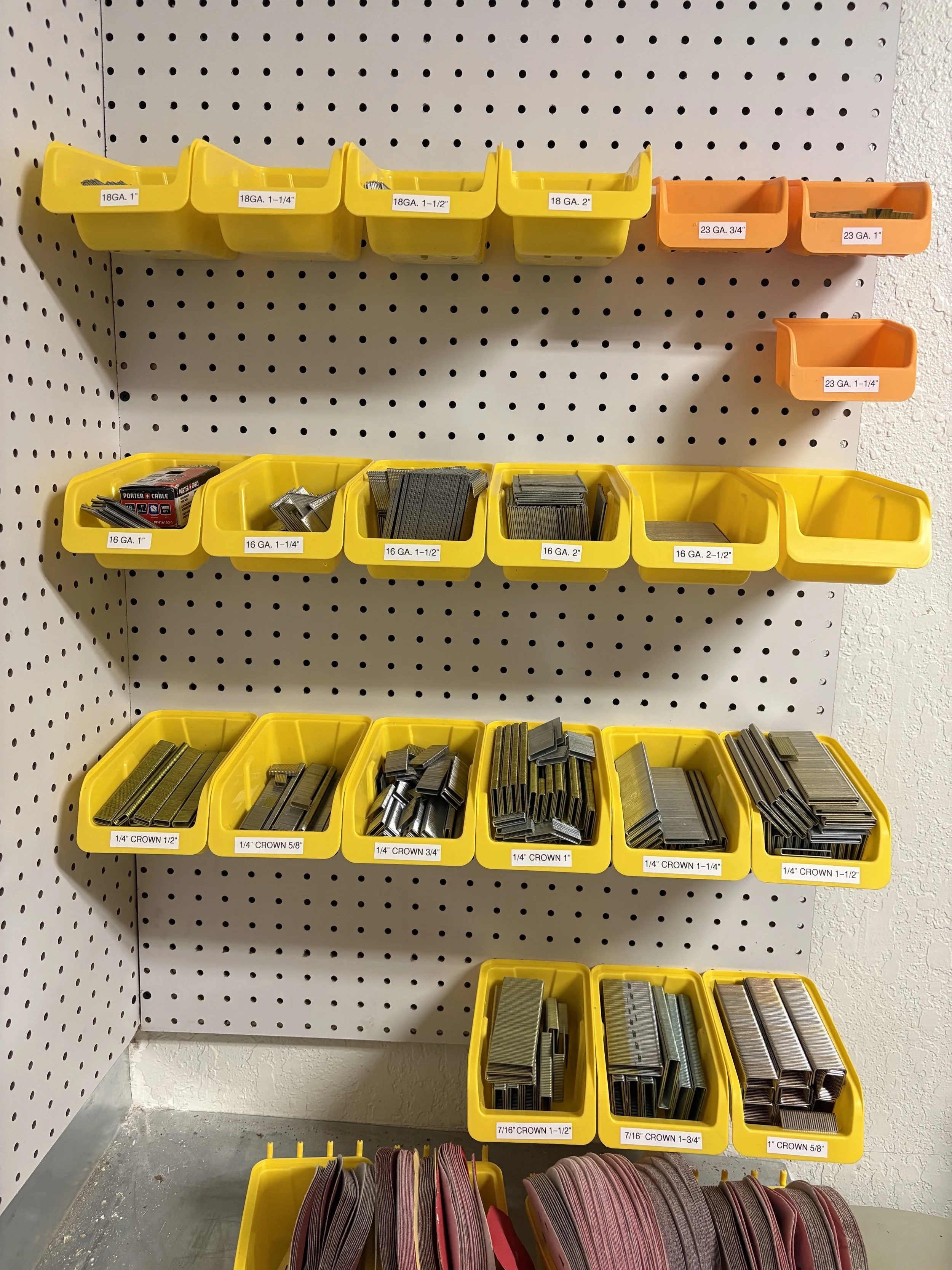 Plastic bins mounted on a pegboard wall containing various hardware supplies, such as nails, screws, and abrasives, with labels indicating sizes and types.