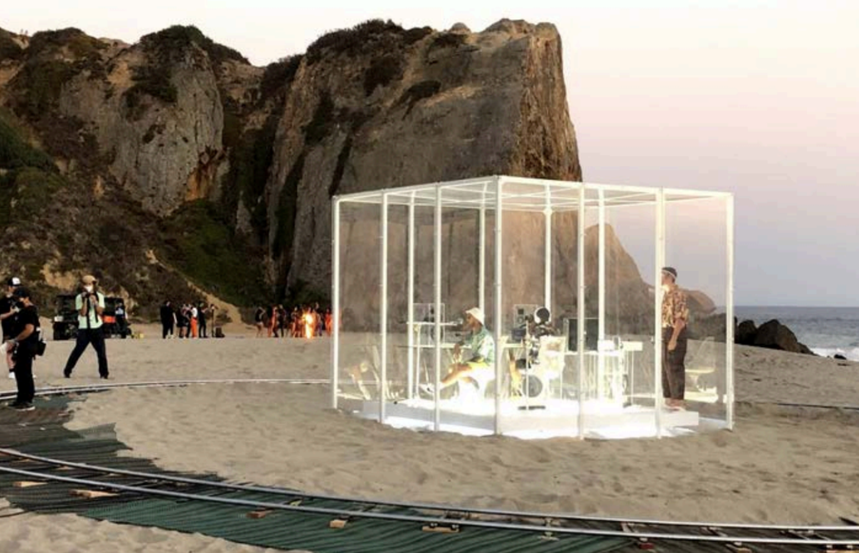 A film crew on a beach with rocky cliffs in the background, with a white transparent structure housing equipment and crew members, while others film or document the scene during sunset.