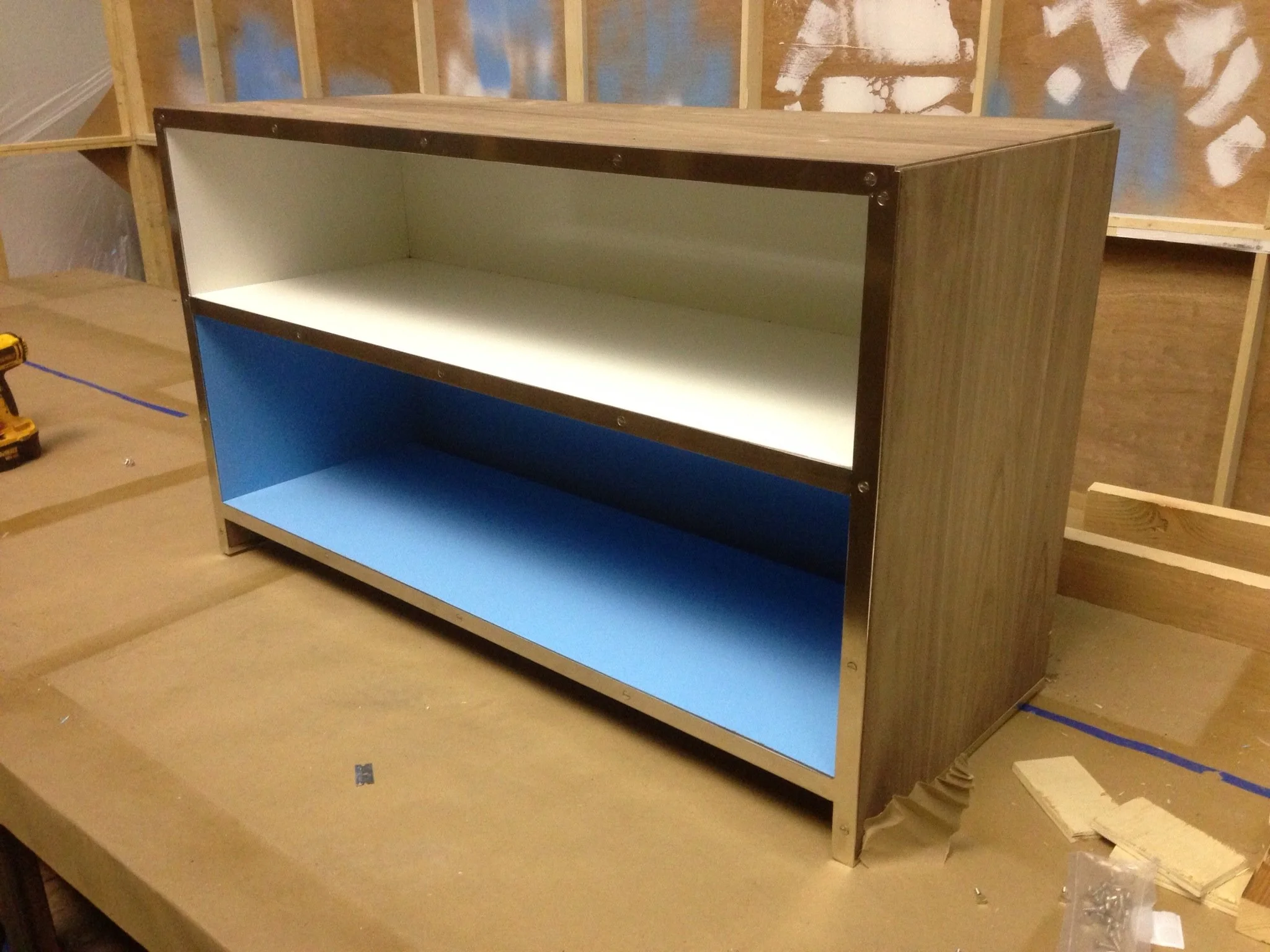New wooden bookshelf with blue backing in a room under construction, surrounded by tools and building materials.