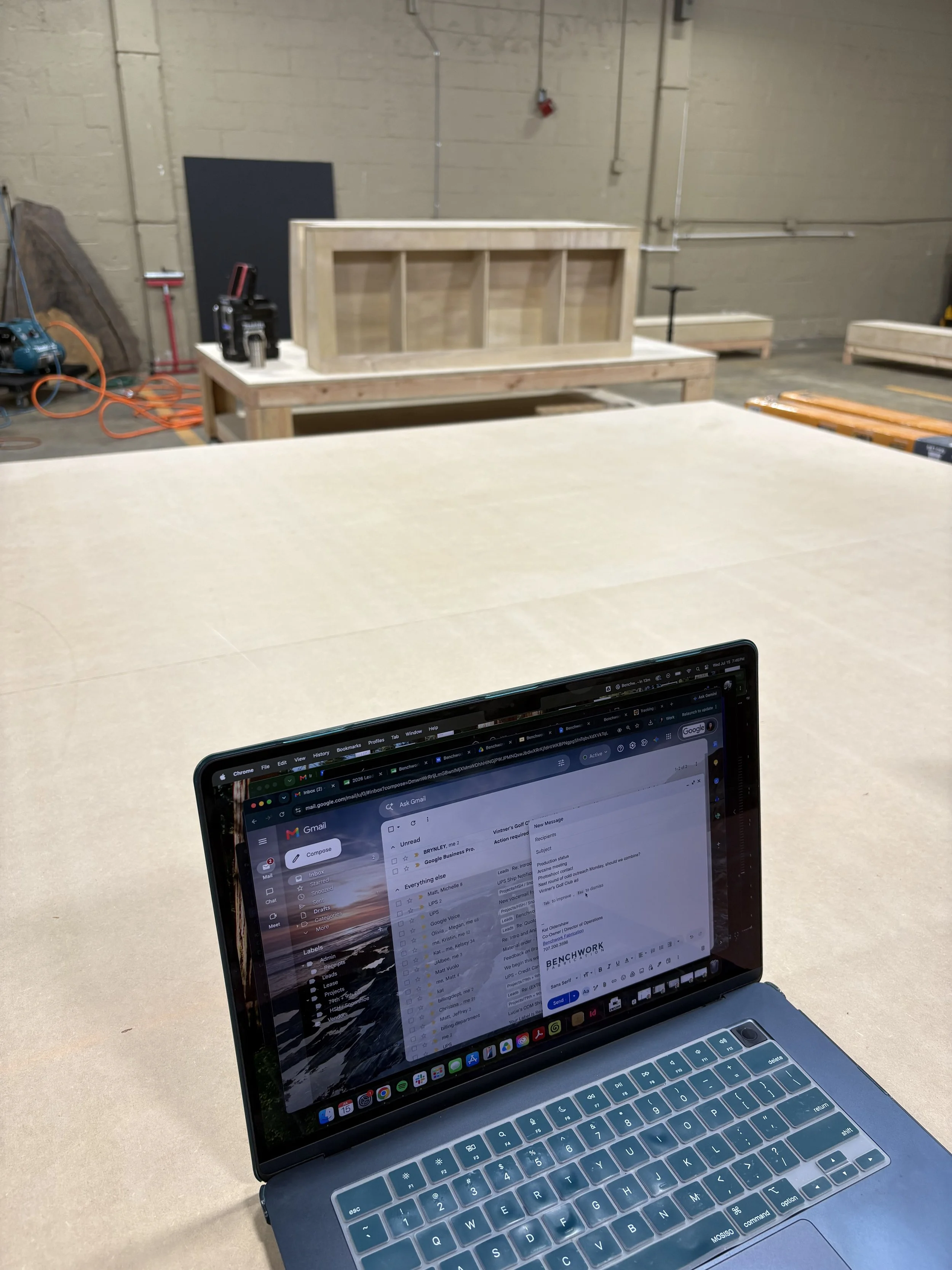 A silver laptop displaying an email inbox on a beige work table in an industrial setting with concrete walls, visible pipework, and construction tools.