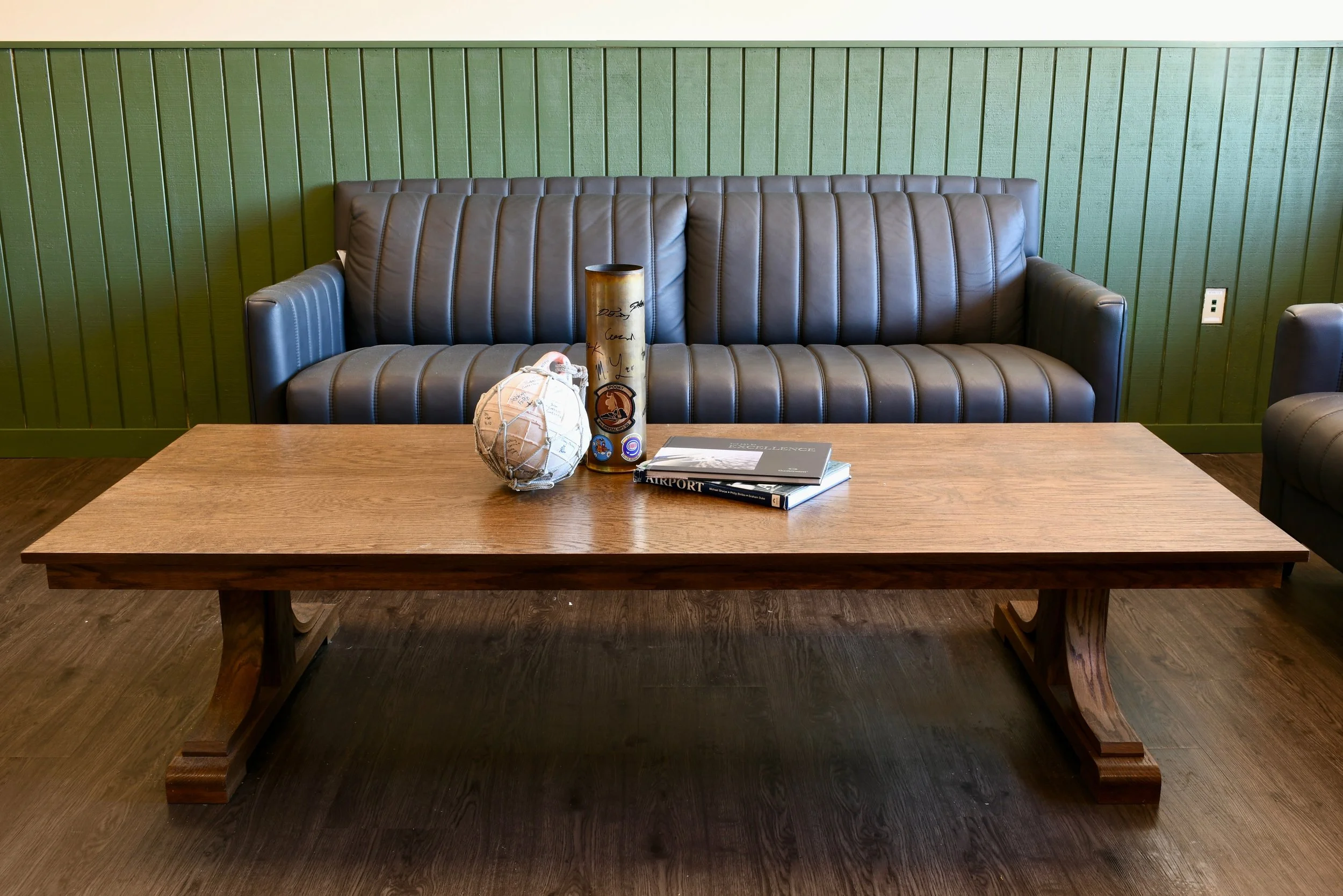 A living room scene with a dark leather sofa with vertical stitching, a wooden coffee table in front, and a green paneled wall behind. On the table are a decorative round object, a tall cylindrical item with signatures, and a small stack of books.