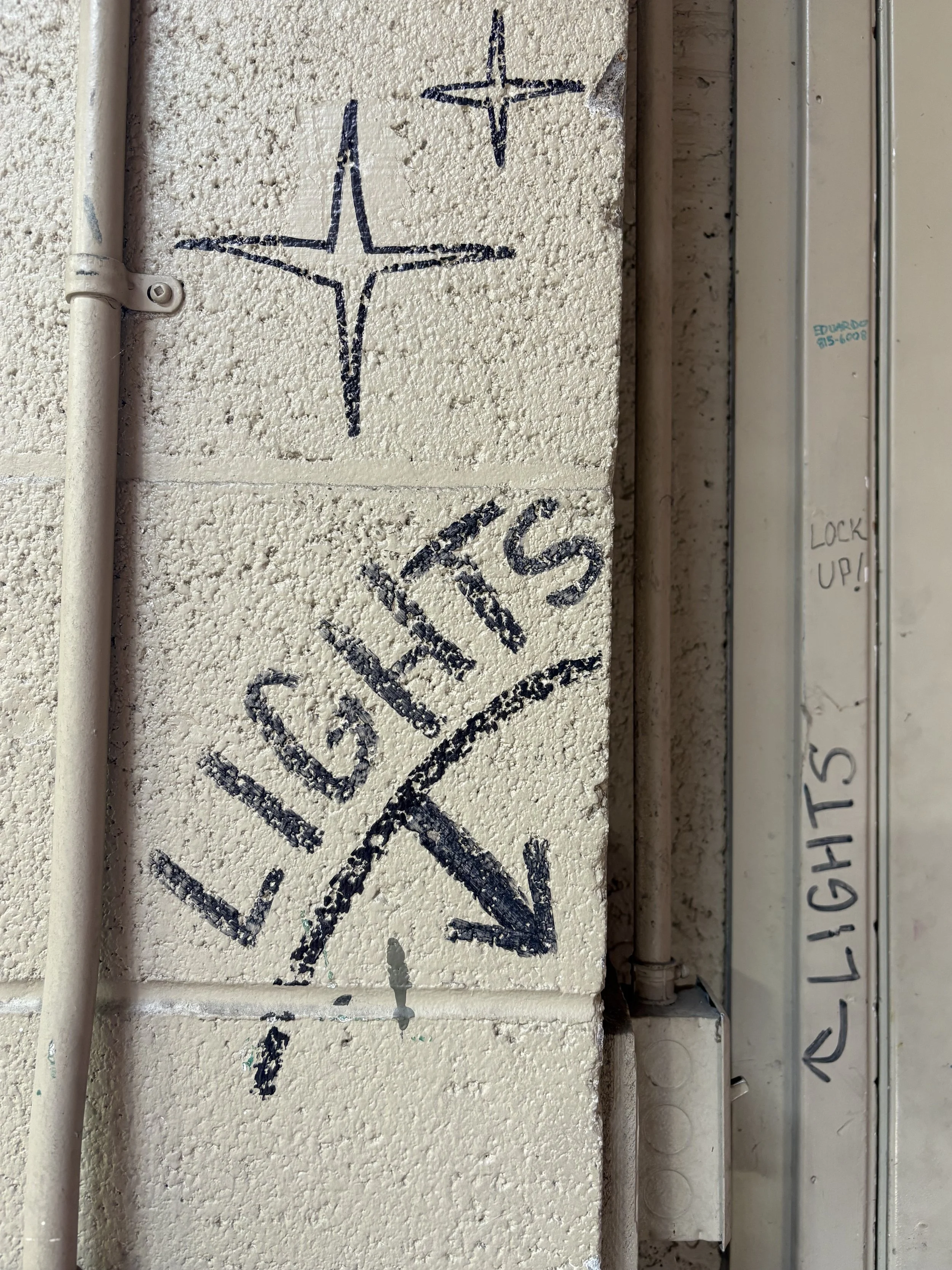 Graffiti of two airplanes drawn with black marker on a textured beige wall, with a white pipe and a metal electrical panel nearby, and part of the words 'LOCK UP!' visible on the panel.