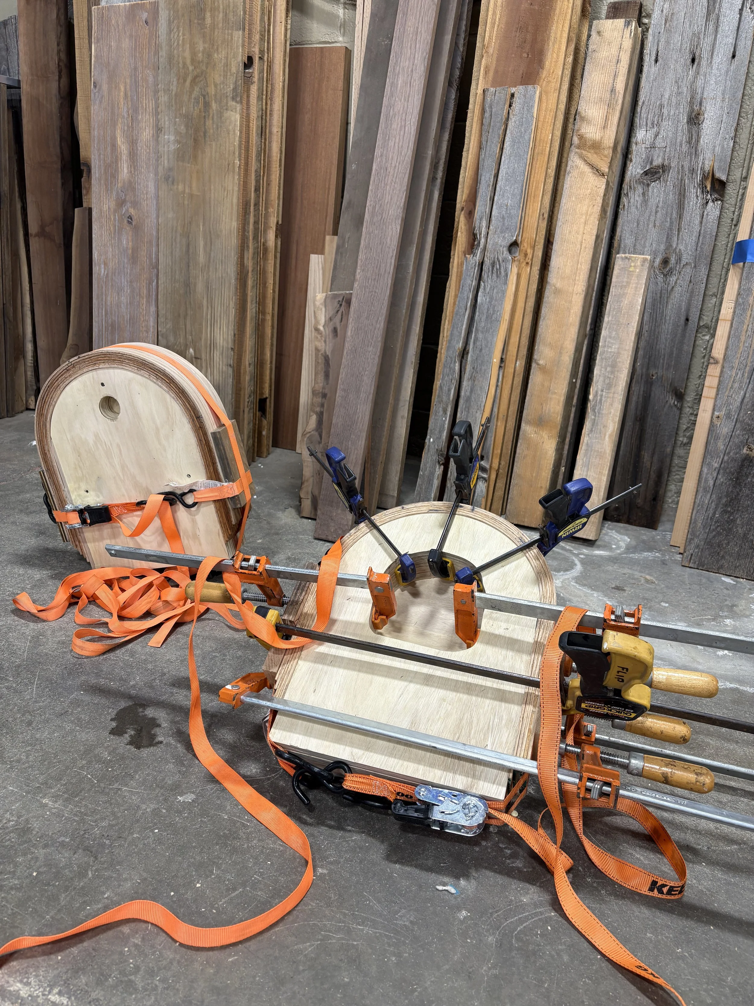 A woodworking shop with various wooden planks leaning against the wall, a clamp on a piece of wood, and orange ratchet straps on the floor.