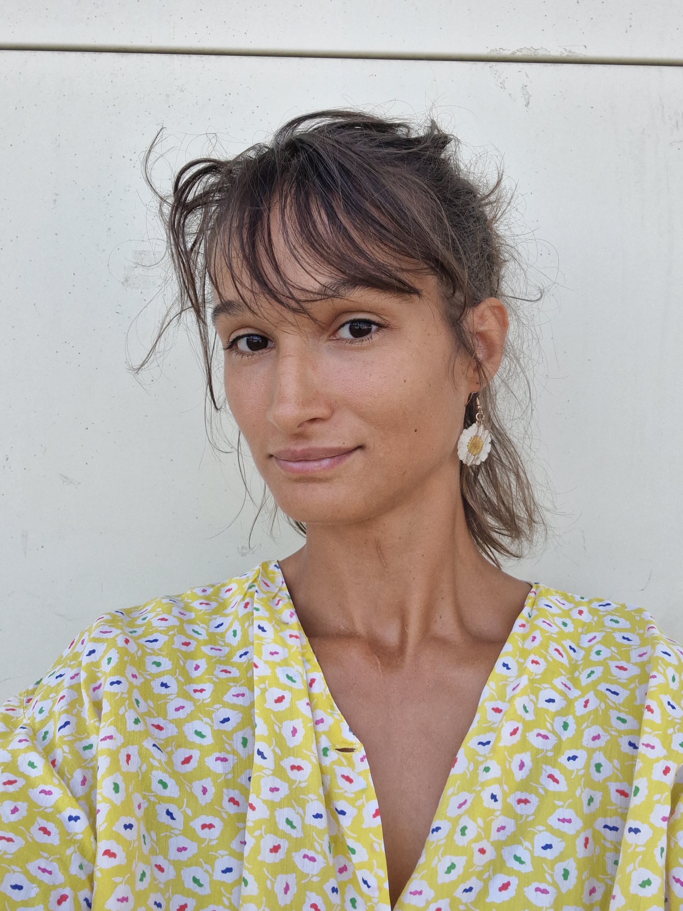 Une femme souriante avec des cheveux courts et ondulés, portant des boucles d'oreilles en forme de marguerite et une robe jaune à motifs floraux.