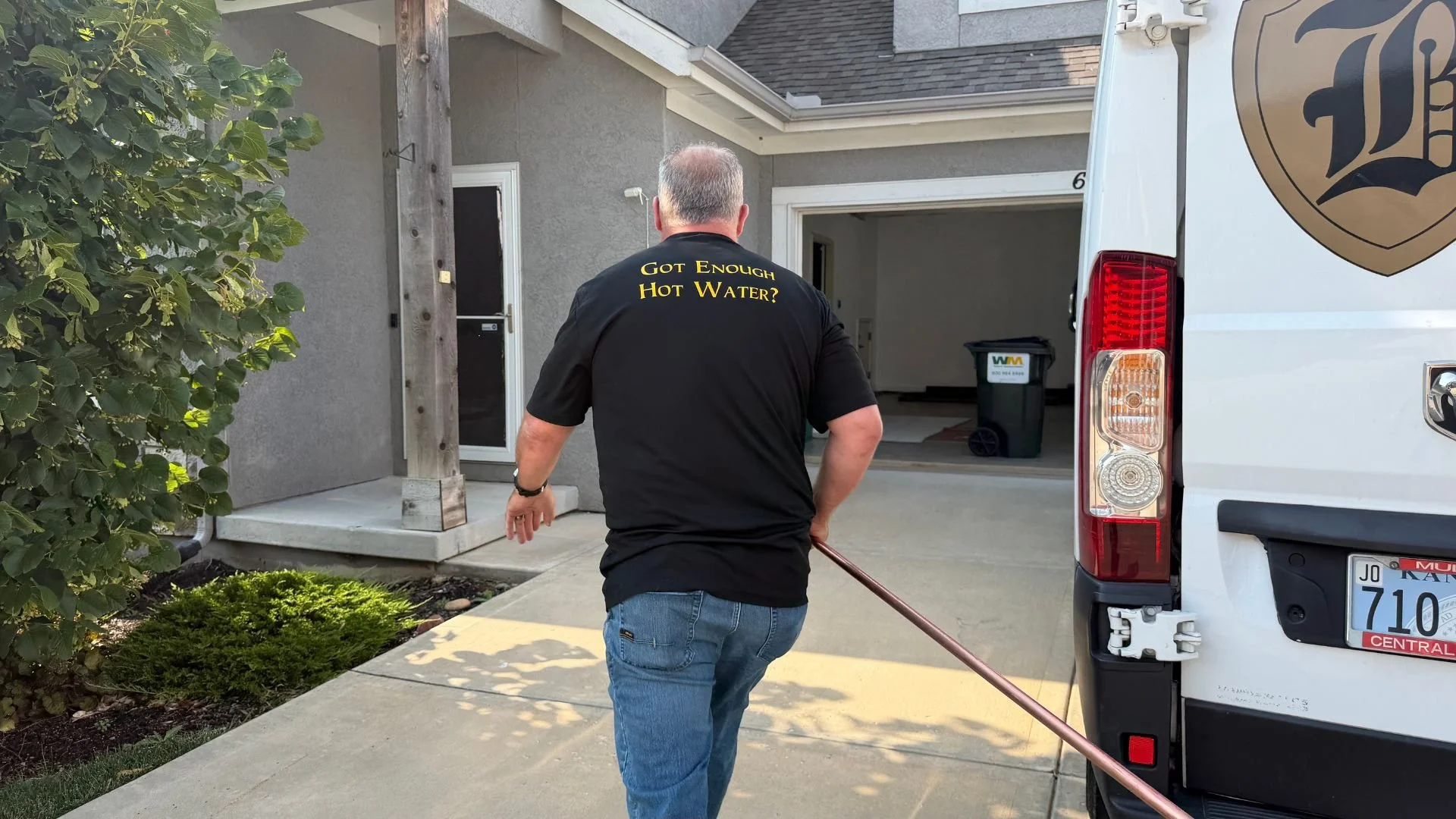 Mike Covington of Bristol Plumbing in front of a Johnson County, Kansas house, wearing a black shirt with the text 'Got Enough Hot Water?' on the back, holding a copper pipe, next to white service van.