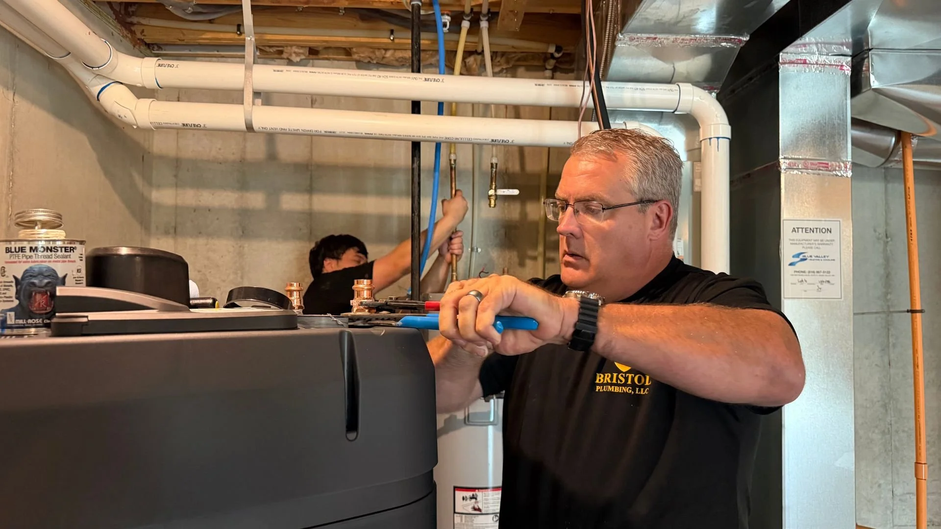 Bristol Plumbing owner Mike Covington working on a black tankless water system while other plumber works on pipes behind him in a Johnson County, Kansas home's basement.