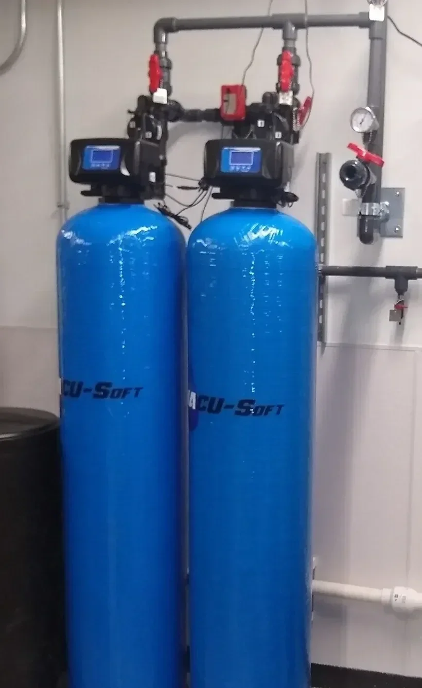 Two large blue cylindrical MAC Water Technologies water softening tanks with digital control heads, connected to black and gray piping and valves mounted on a wall.