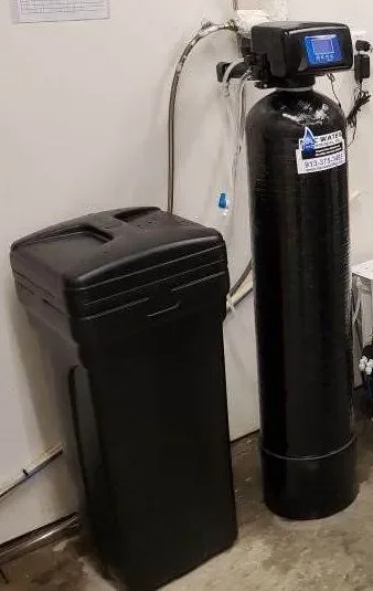 A water softening system with a black softener tank and a black cylinder unit next to it connected to wall of a utility room in a Kansas City home.
