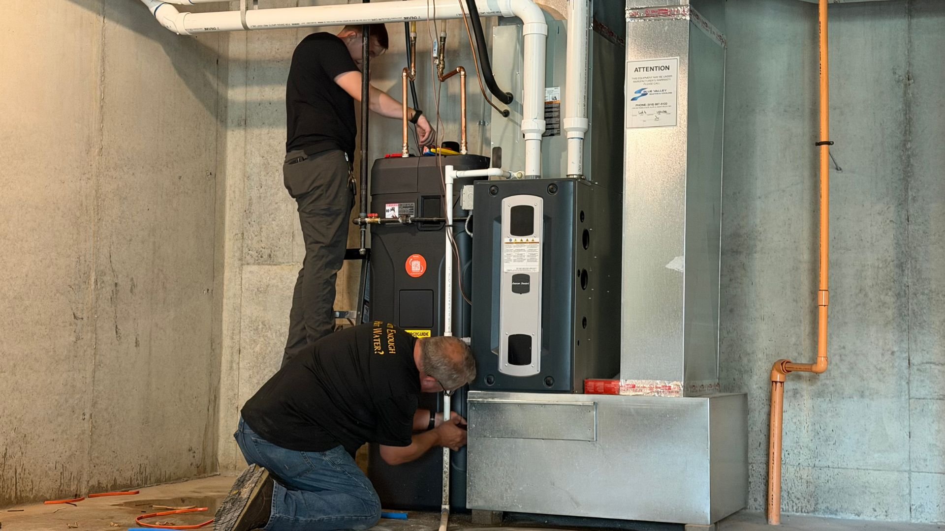 Two technicians working on an Essency water heater system, with one kneeling and the other standing, surrounded by pipes and electrical components.
