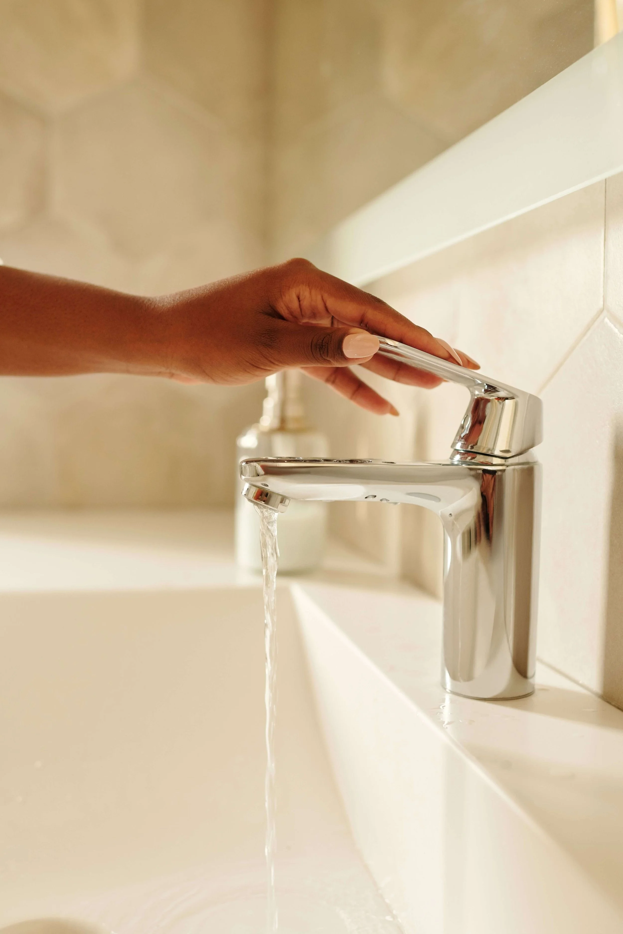 Close-up of a hand turning on a modern chrome faucet with water flowing into a sink, in a bathroom or washbasin area.