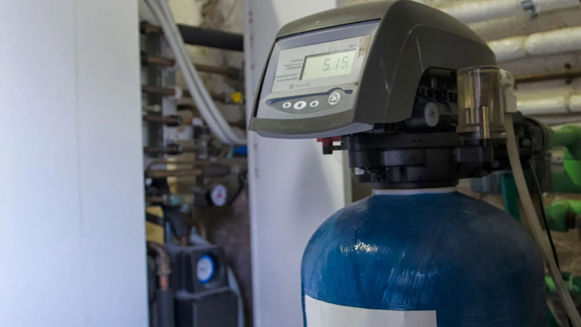 A digital control panel attached to a water softener cylindrical tank in a utility space.