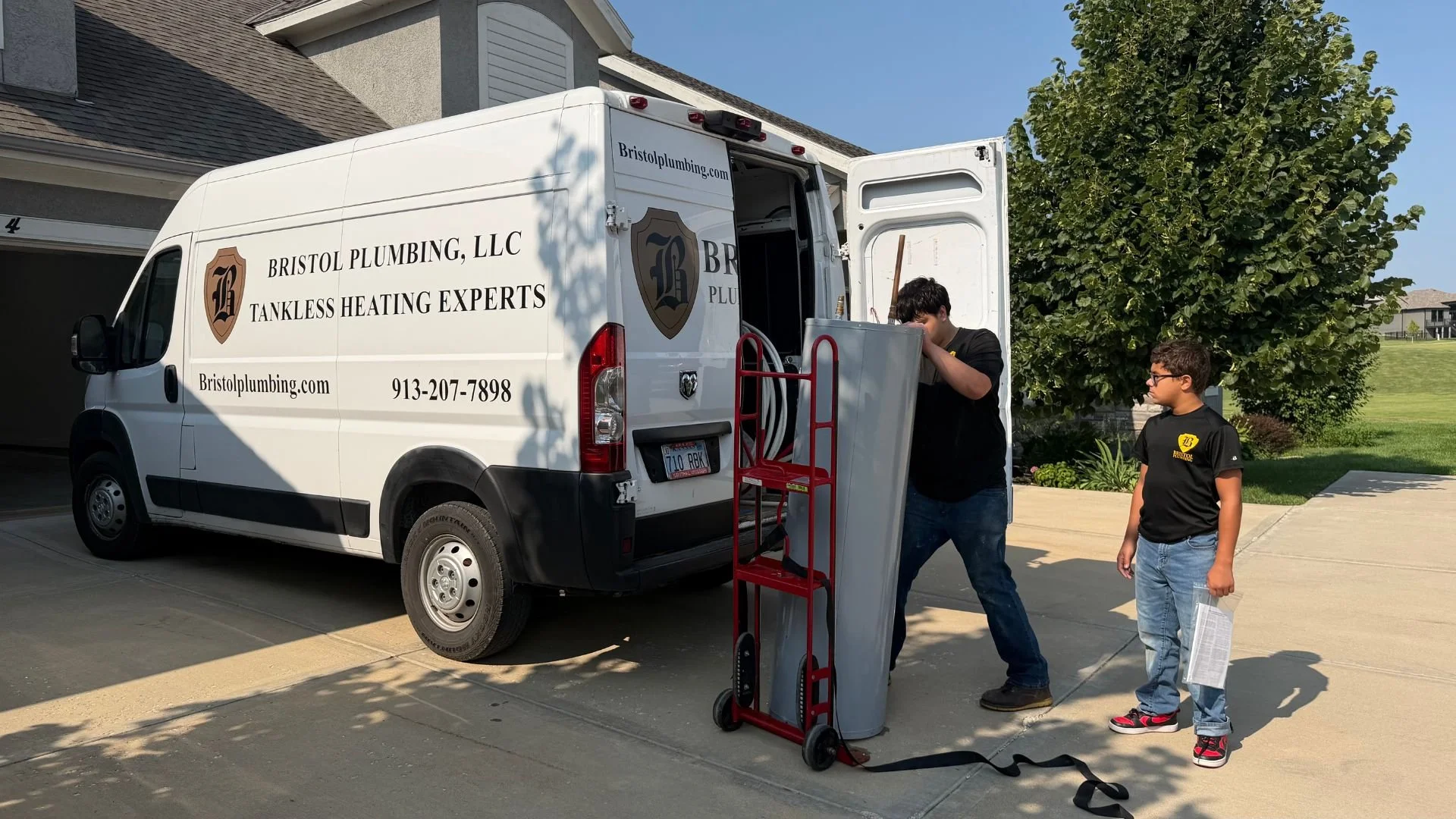 Two young men loading equipment into a white plumbing van parked in a driveway. The van has the logo and contact information for Bristol Plumbing, LLC, a tankless heating expert.