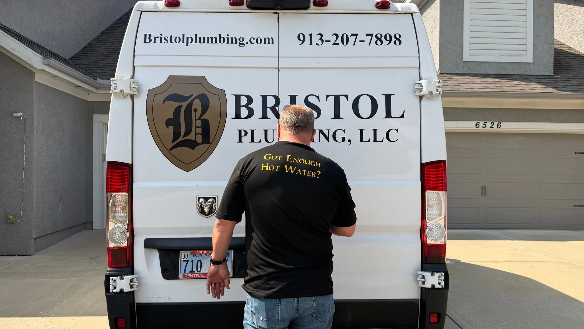Mike Covington standing behind a white Bristol Plumbing, LLC service vehicle, wearing a black shirt with the text 'Got Enough Hot Water?' on the back, parked in front of a residential house.