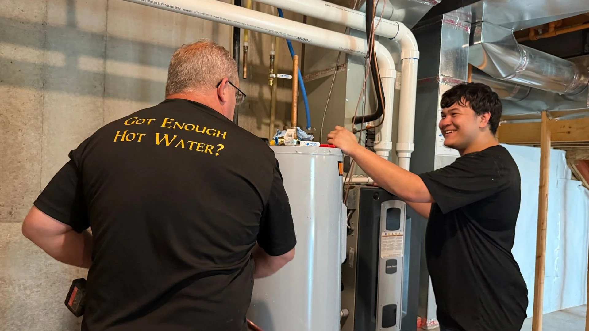 Bristol Plumbing owner Mike Covington wearing black shirt that reads 'Got Enough Hot Water?' and another plumber who is smiling working on tankless water heater in a Kansas City home's basement.