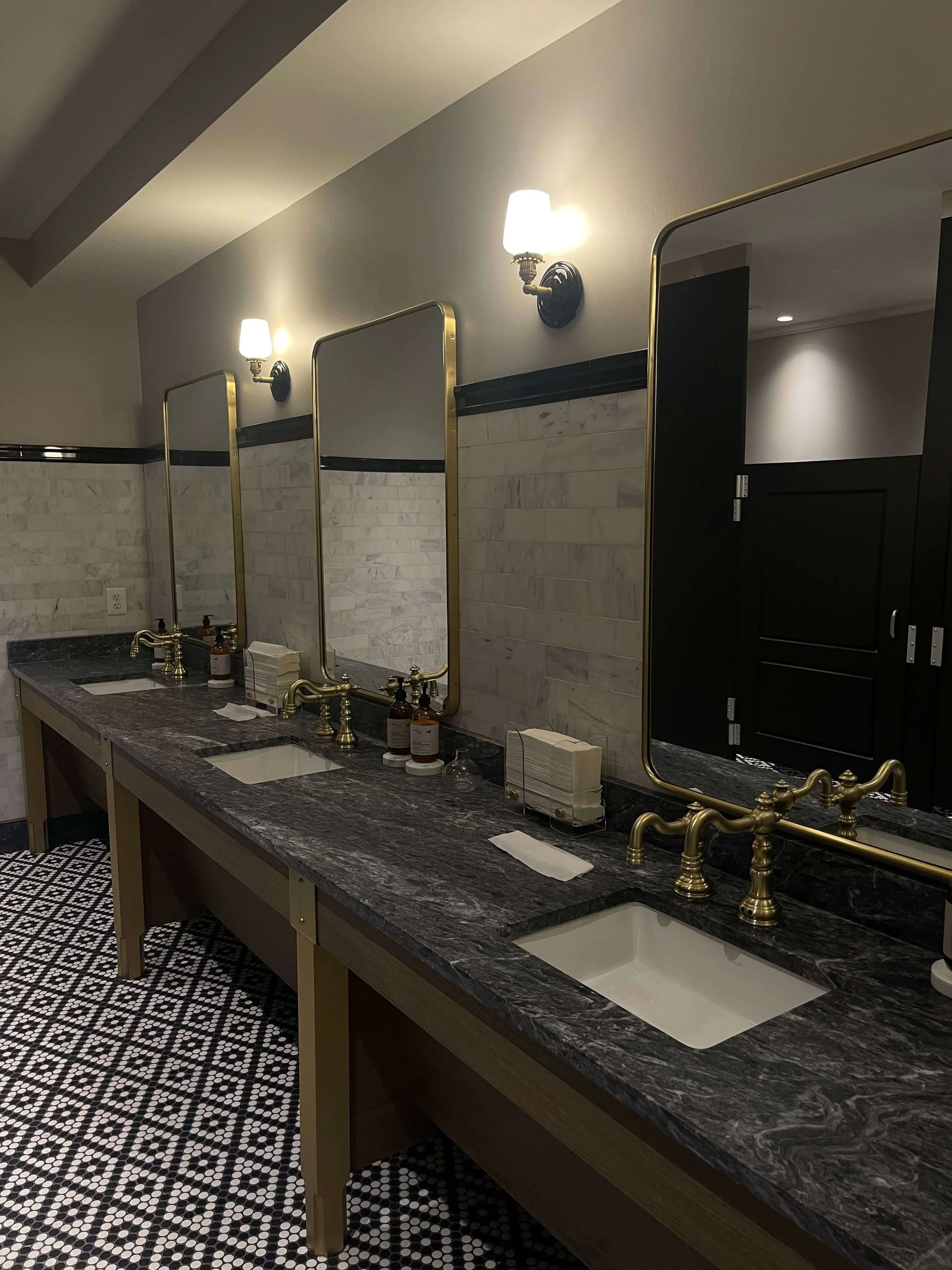 Elegant public bathroom in Kansas City with dark patterned floor, marble countertop with two sinks, and gold fixtures.