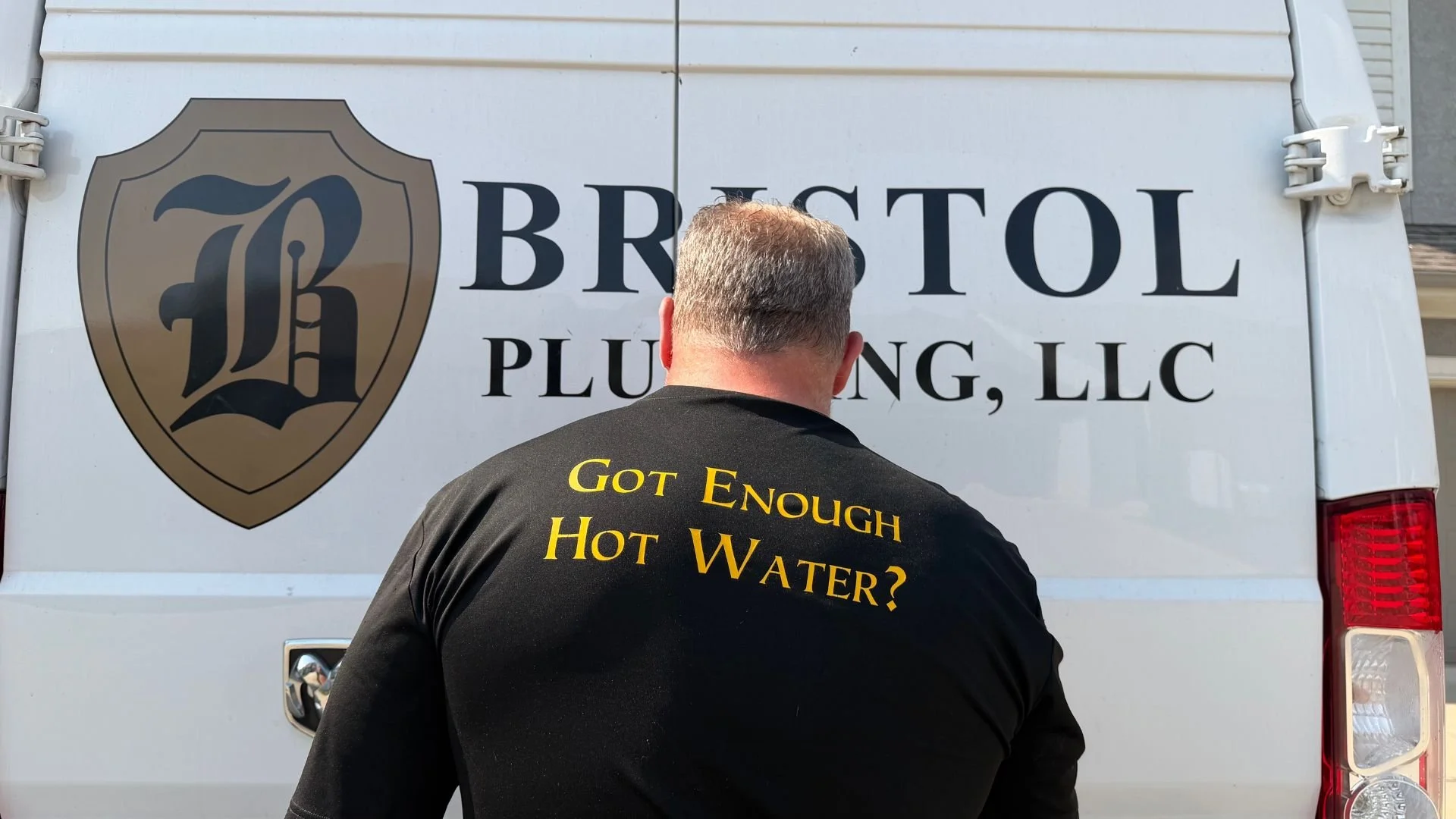 Mike Covington standing behind a white Bristol Plumbing, LLC service vehicle, wearing a black shirt with the text 'Got Enough Hot Water?' on the back, parked in front of a residential house.
