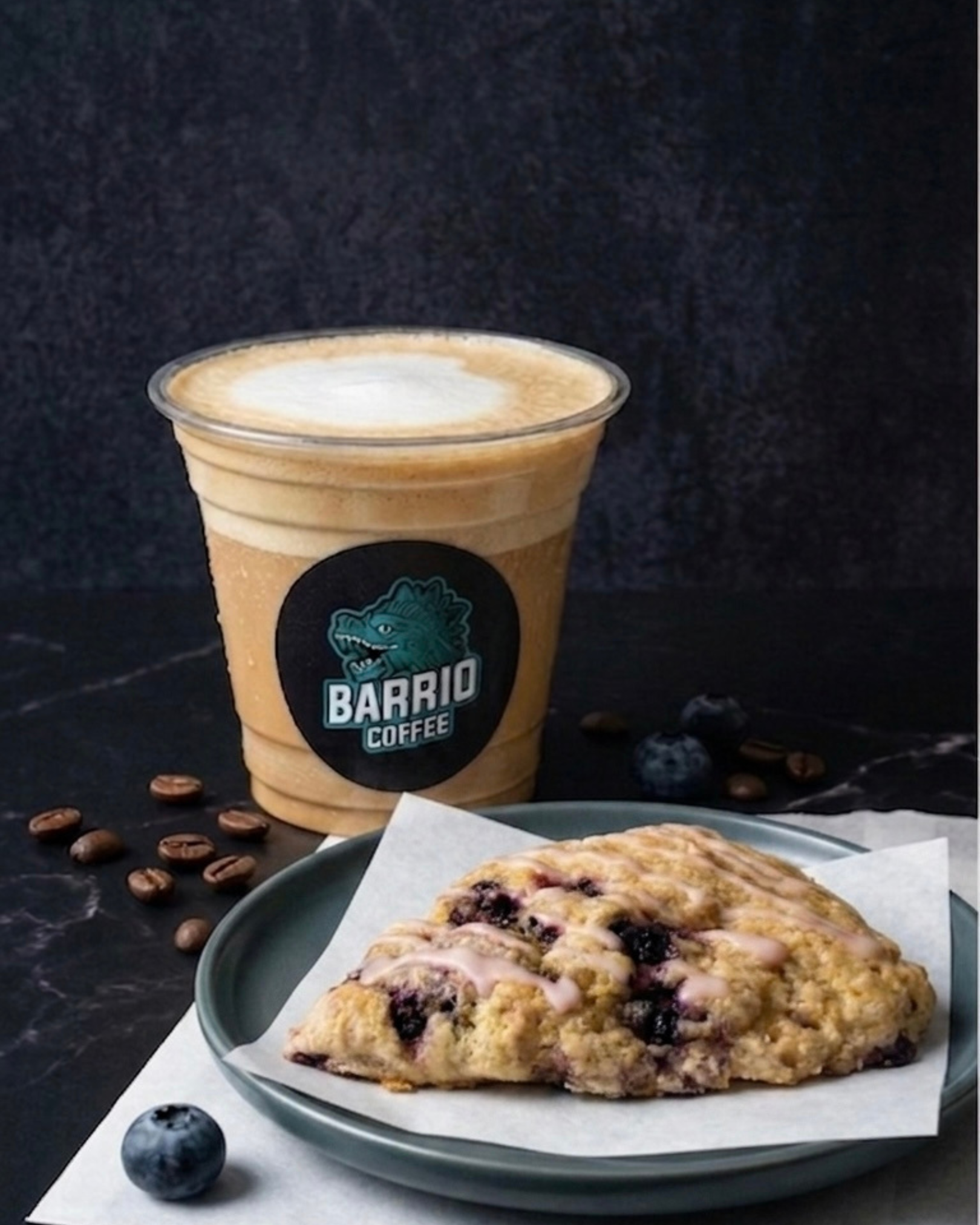 A blueberry scone with blueberries and icing on a white paper, a bowl with blueberry dessert and a cup of coffee with a logo and a wolf, coffee beans scattered on a black background.
