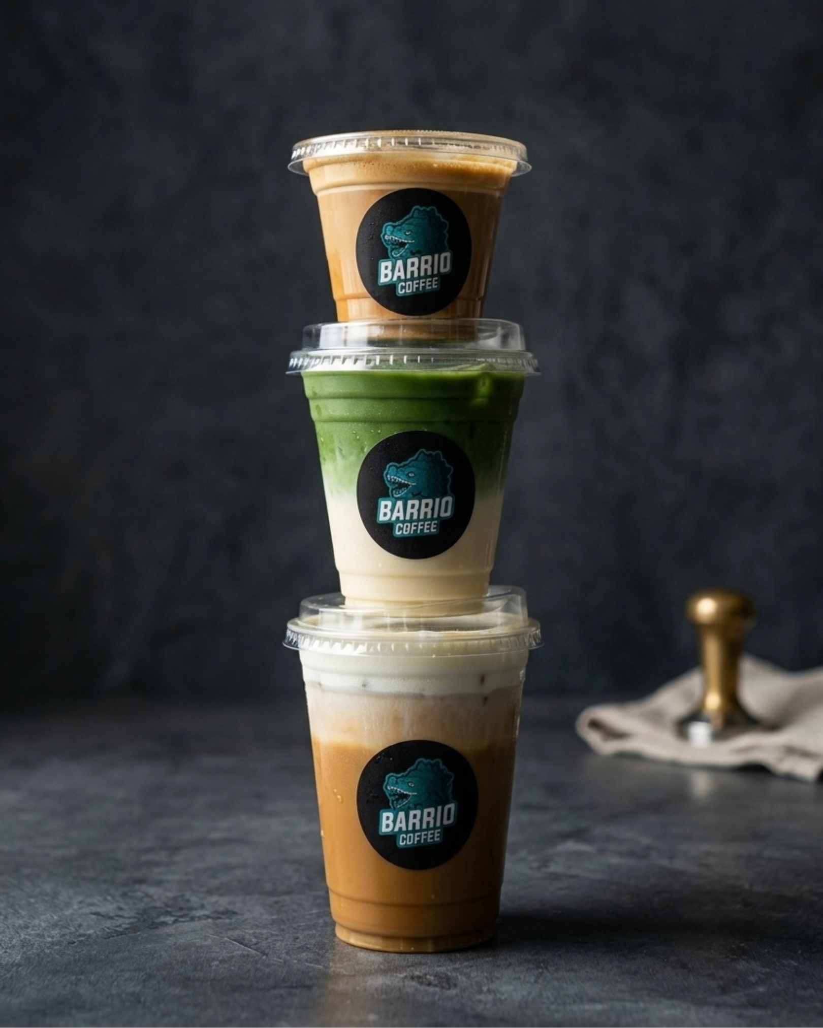 Stacked iced coffee drinks in clear plastic cups with black and teal Barrio Coffee labels, set against a dark background.