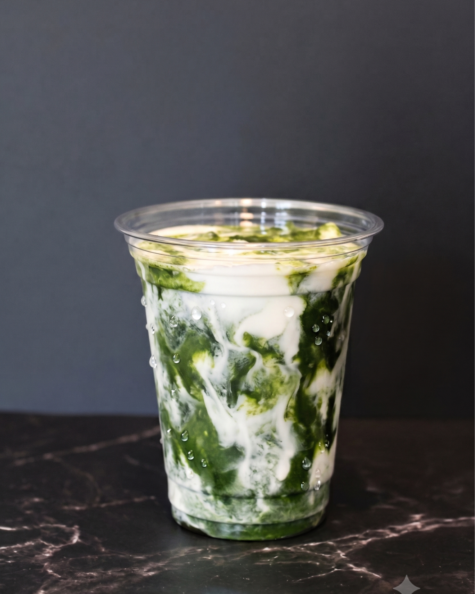 A clear plastic cup filled with a layered green and white dessert, possibly matcha and cream, set on a dark marble surface with a black background.