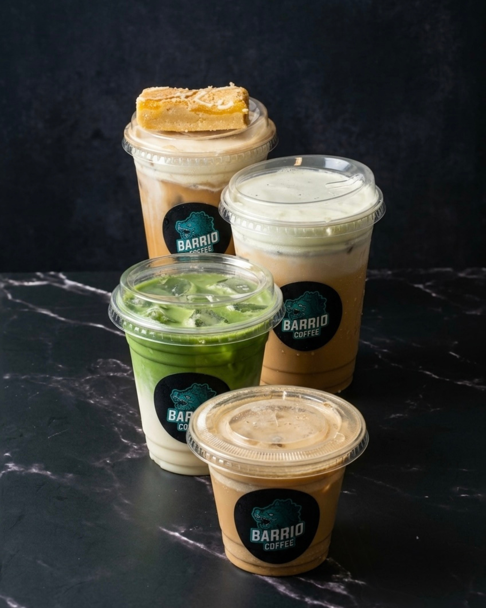 Multiple cold coffee drinks in clear plastic cups with black and teal Barrio Coffee logos, one with a cookie on top, another with whipped cream, and others with different toppings, all set against a dark textured background.