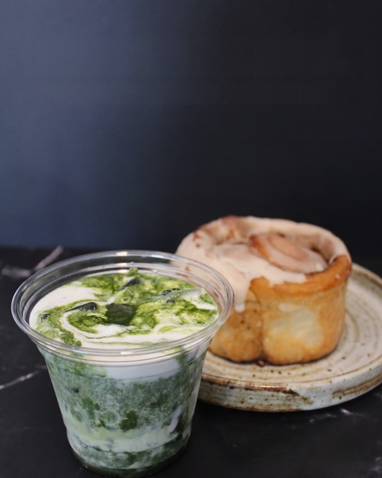 A clear plastic cup filled with a green and white swirl of a creamy drink, placed on a black surface. Behind it, a plate with a baked item topped with a topping, possibly meat or cheese.