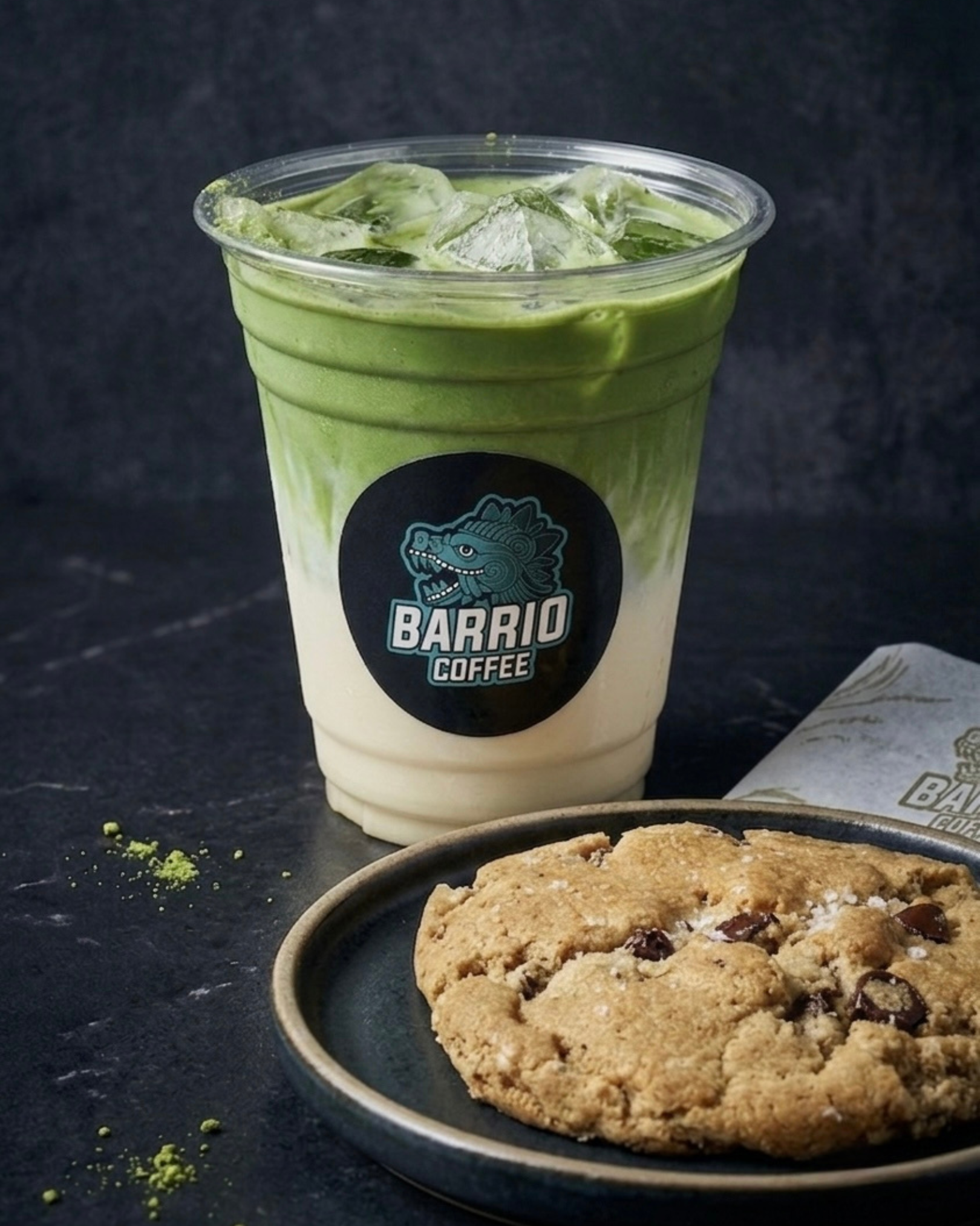 A clear plastic cup with a logo for Barrio Coffee contains a green iced matcha latte with ice cubes. In the foreground, there's a bowl of chocolate chip cookies on a dark surface, with some green matcha powder and crumbs scattered around.