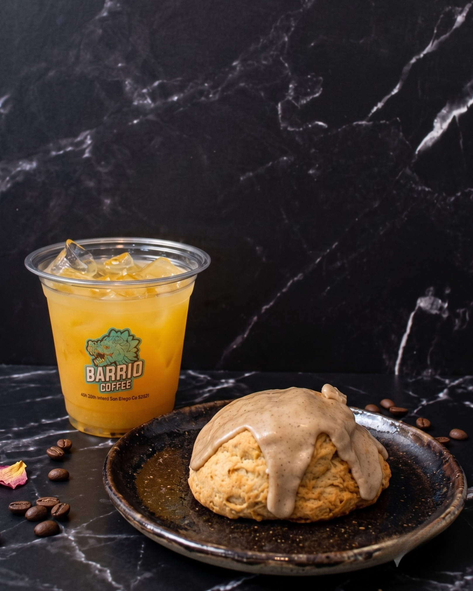 A black plate with a large cookie topped with ice cream and caramel sauce, set on a dark surface with scattered coffee beans, alongside a cup of orange-colored drink with ice cubes from Barrio Coffee, with a black marble background.