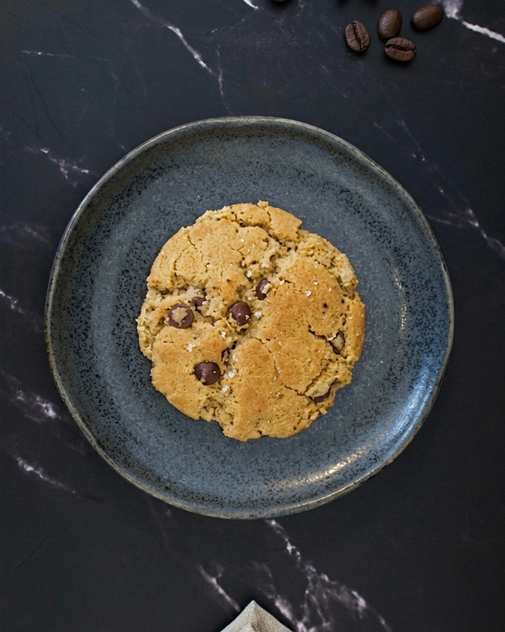 A chocolate chip cookie on a dark, textured plate with coffee beans and salt sprinkled on top. The plate is on a black marble surface with a napkin in the bottom corner.