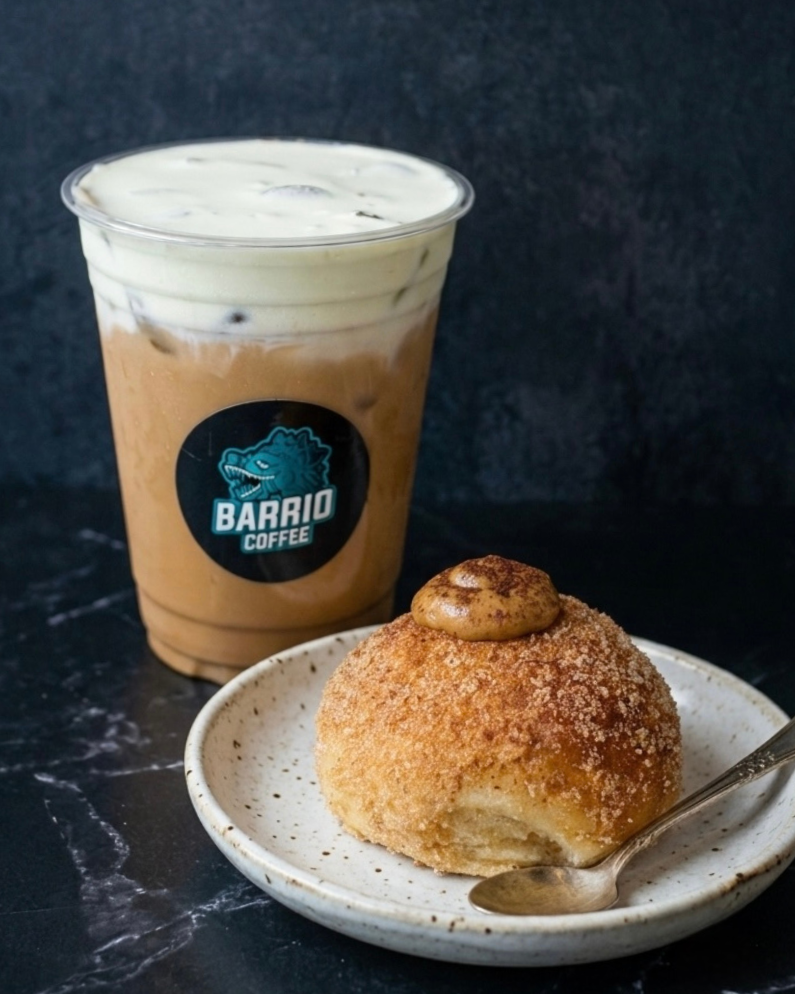 A cup of iced coffee with whipped cream on top and a cookie with a dollop of nut butter on a white speckled bowl with a spoon, on a dark marble countertop.