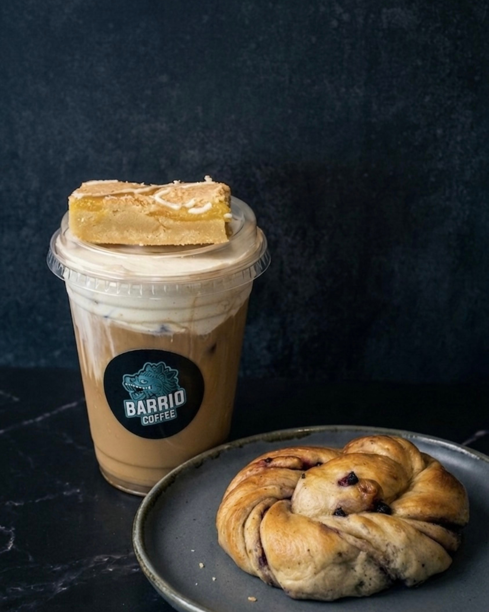 Close-up of a beverage in a plastic cup with a black logo reading 'BARRIO COFFEE' and a pastry with fruit filling on a plate, set against a dark background.