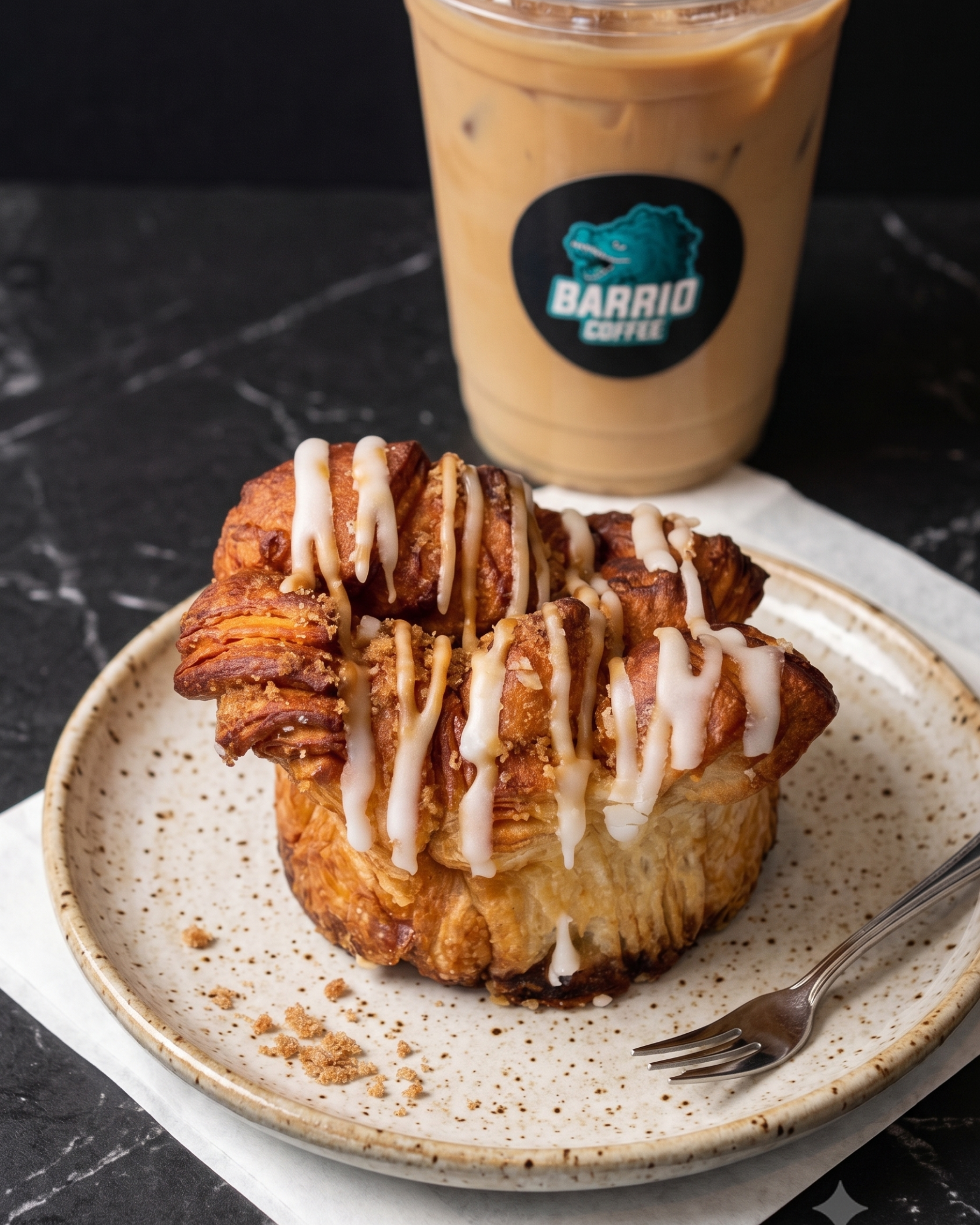 A plate with a fried dessert topped with white icing drizzle, served with a fork, and a cup of iced coffee in the background with a logo that says Barrio Coffee.