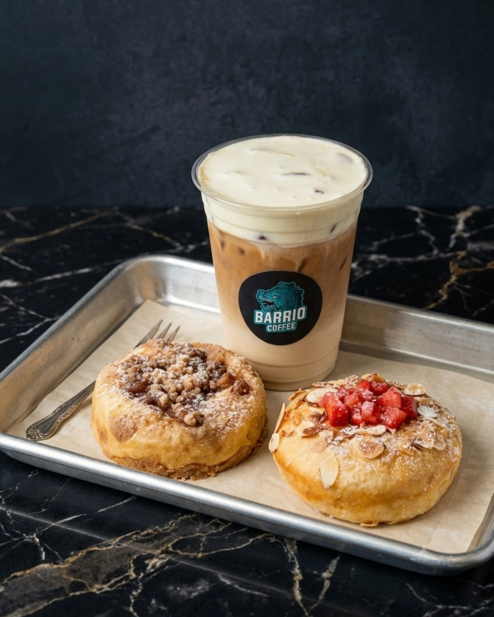A tray with two glazed donuts, one topped with crumbs and chocolate, the other with strawberries and almonds, and a cup of iced coffee with cream at a cafe.