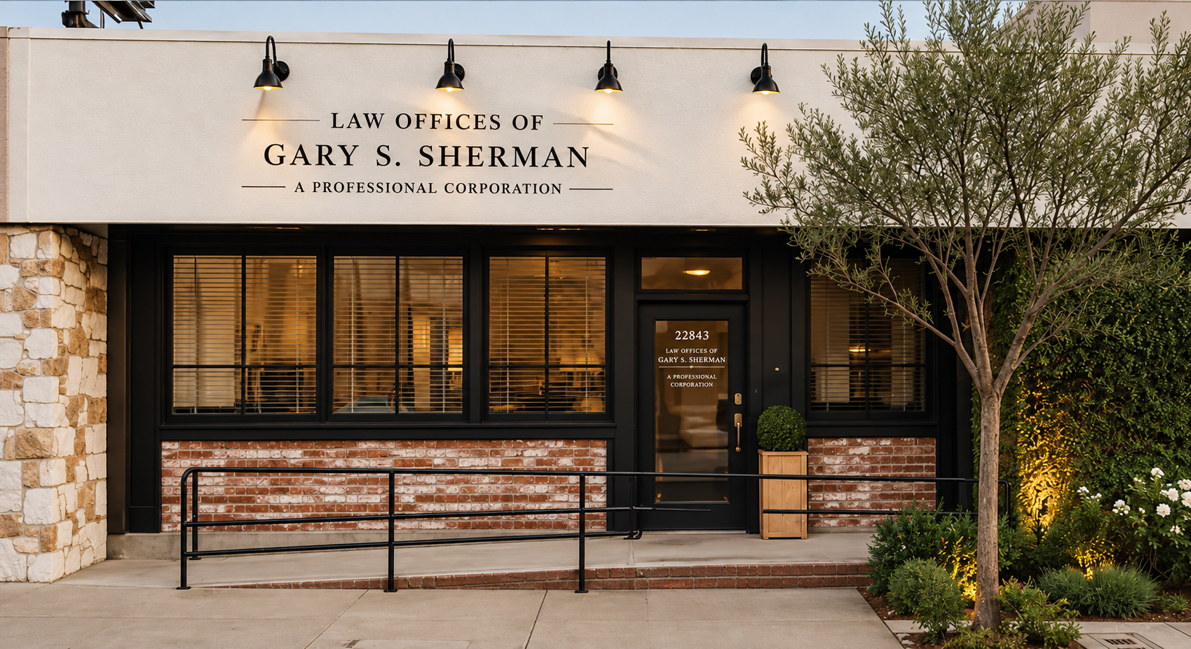 Exterior of a law office building with a sign reading "Law Offices of Gary S. Sherman, A Professional Corporation." The building has large black-framed windows, brick and stone accents, potted plants, a small tree, and exterior lighting.