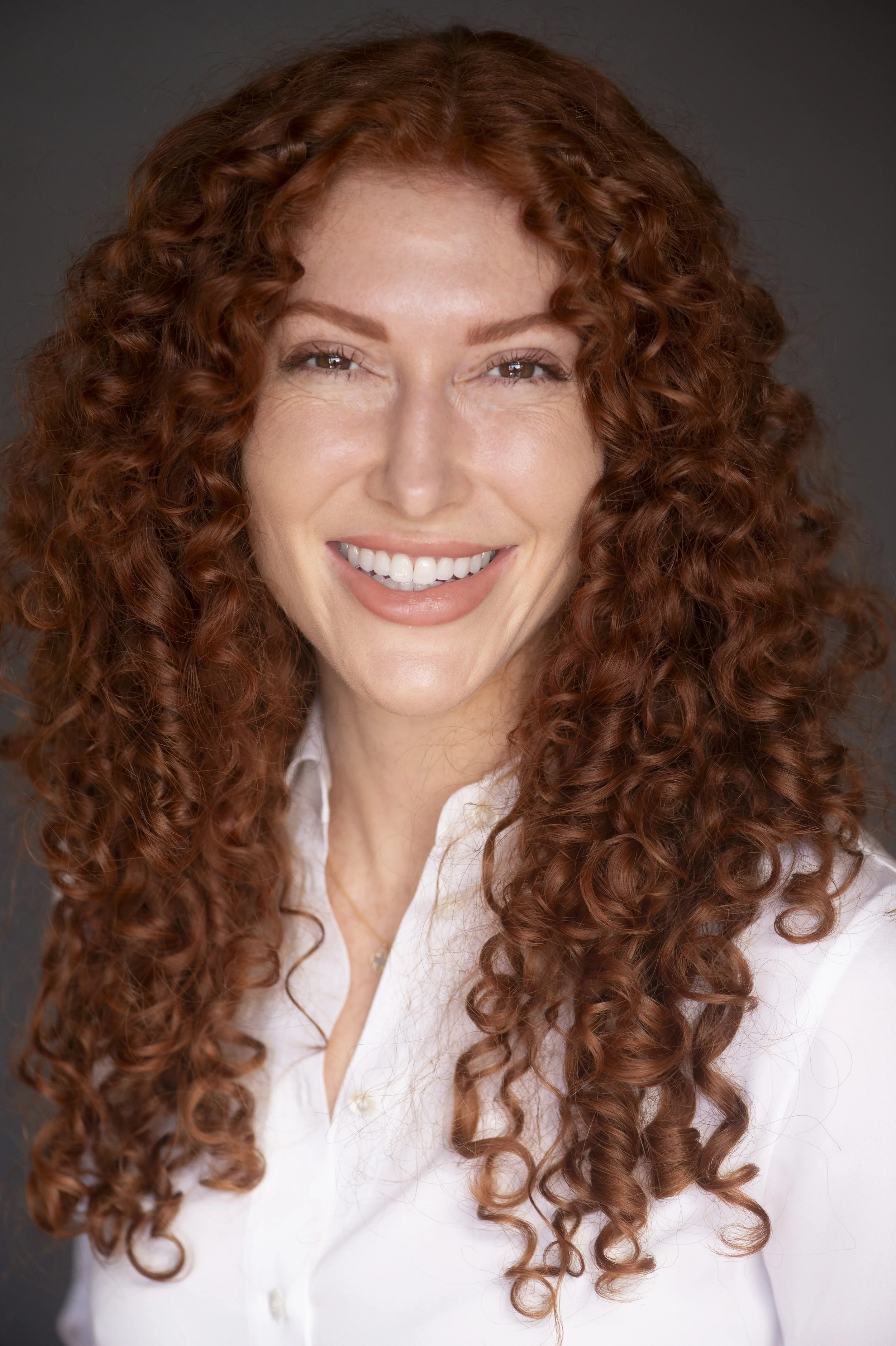 Close-up of a smiling Marlee Elise Sherman, lawyer in Wooland Hills with long, curly red hair, wearing a white shirt, against a dark background.