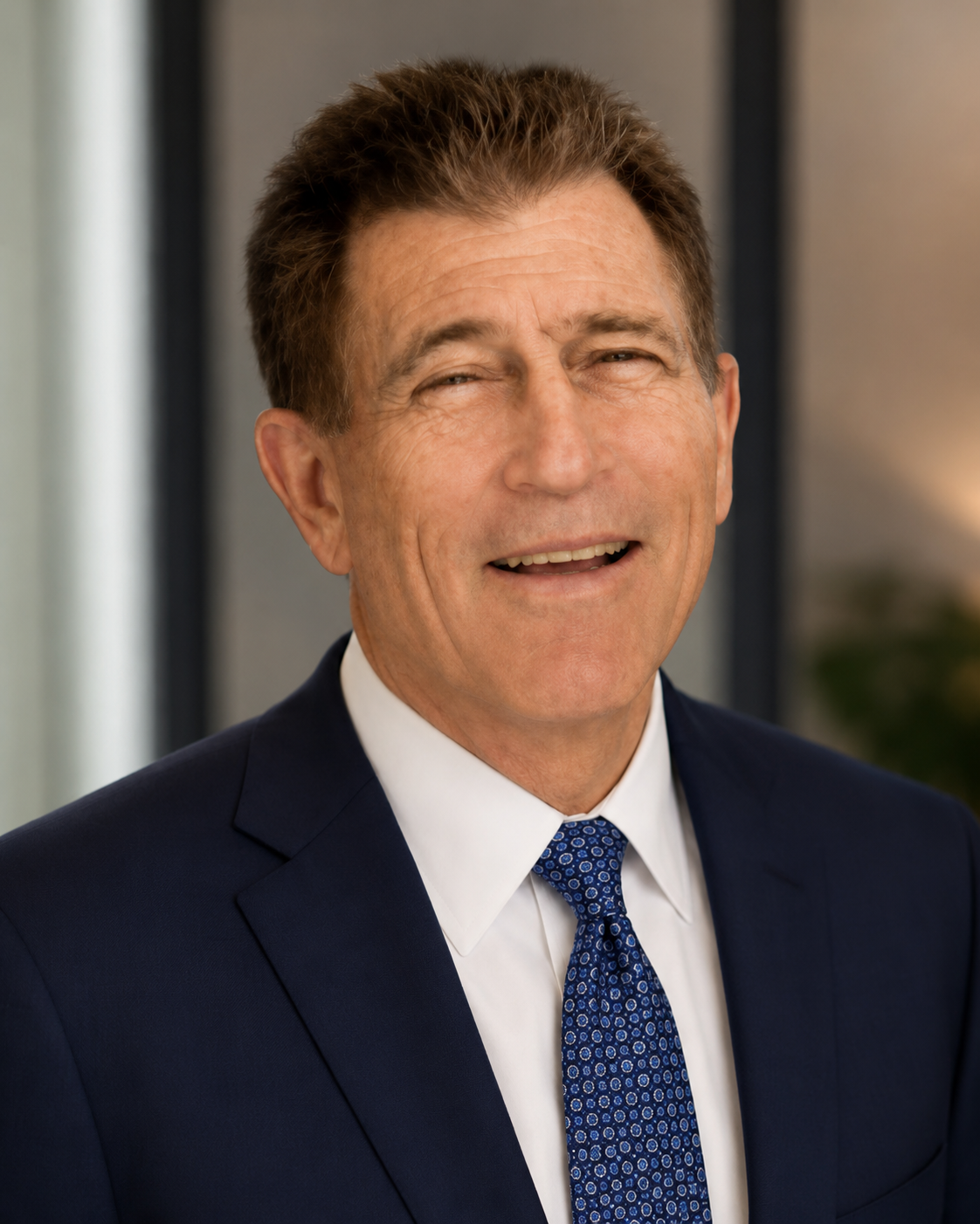 Close-up of a smiling Gary Sherman, lawyer in Woodland Hills with brown hair, wearing a navy suit, white shirt, and blue patterned tie, standing indoors.