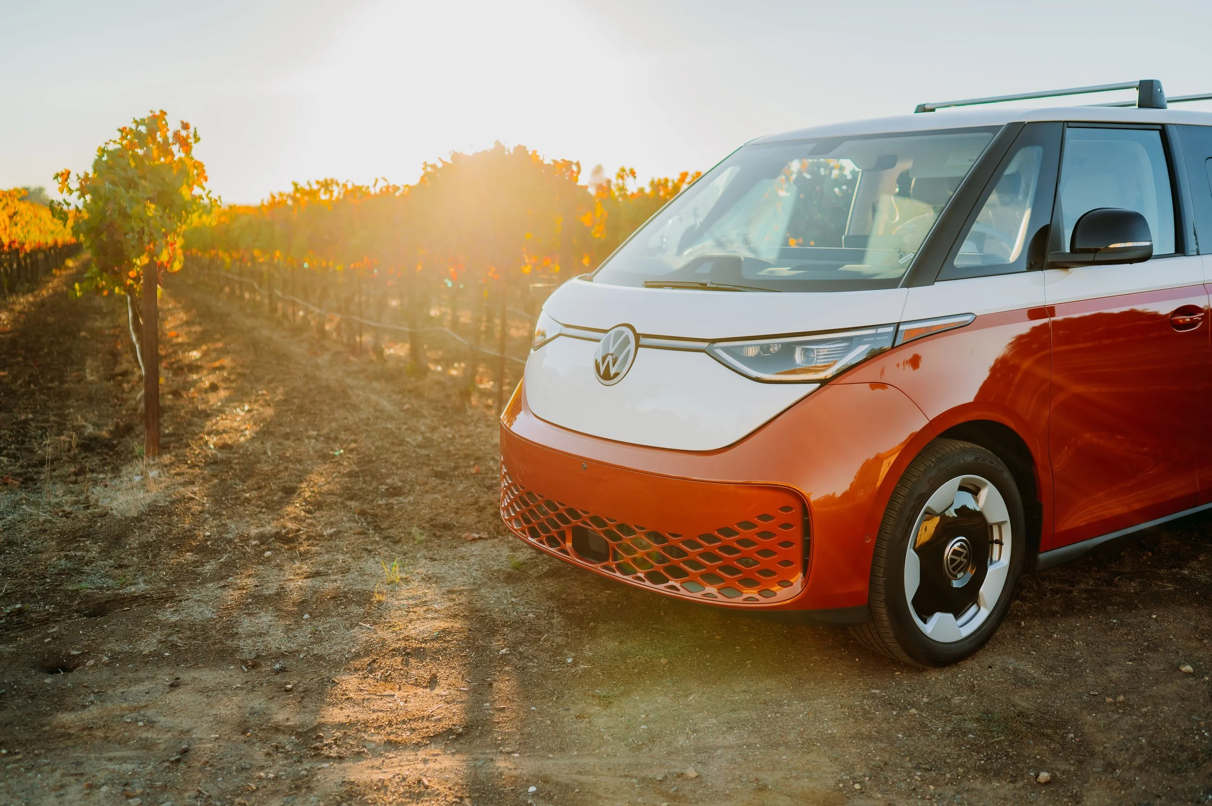 An orange and white Volkswagen electric vehicle parked on a dirt path in a vineyard at sunset.