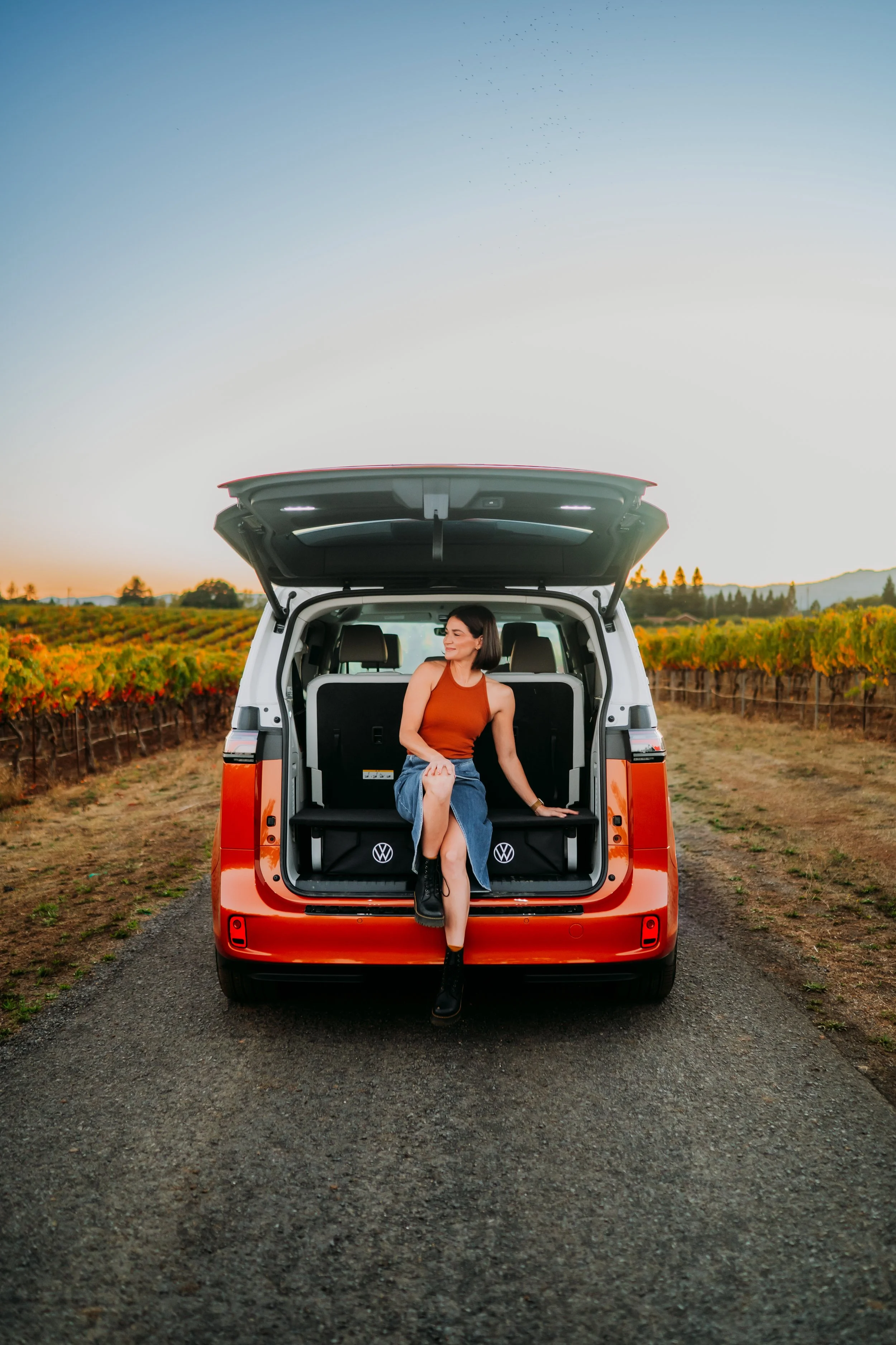 A woman sitting in the back of an orange SUV with the hatch open, in a vineyard during sunset.