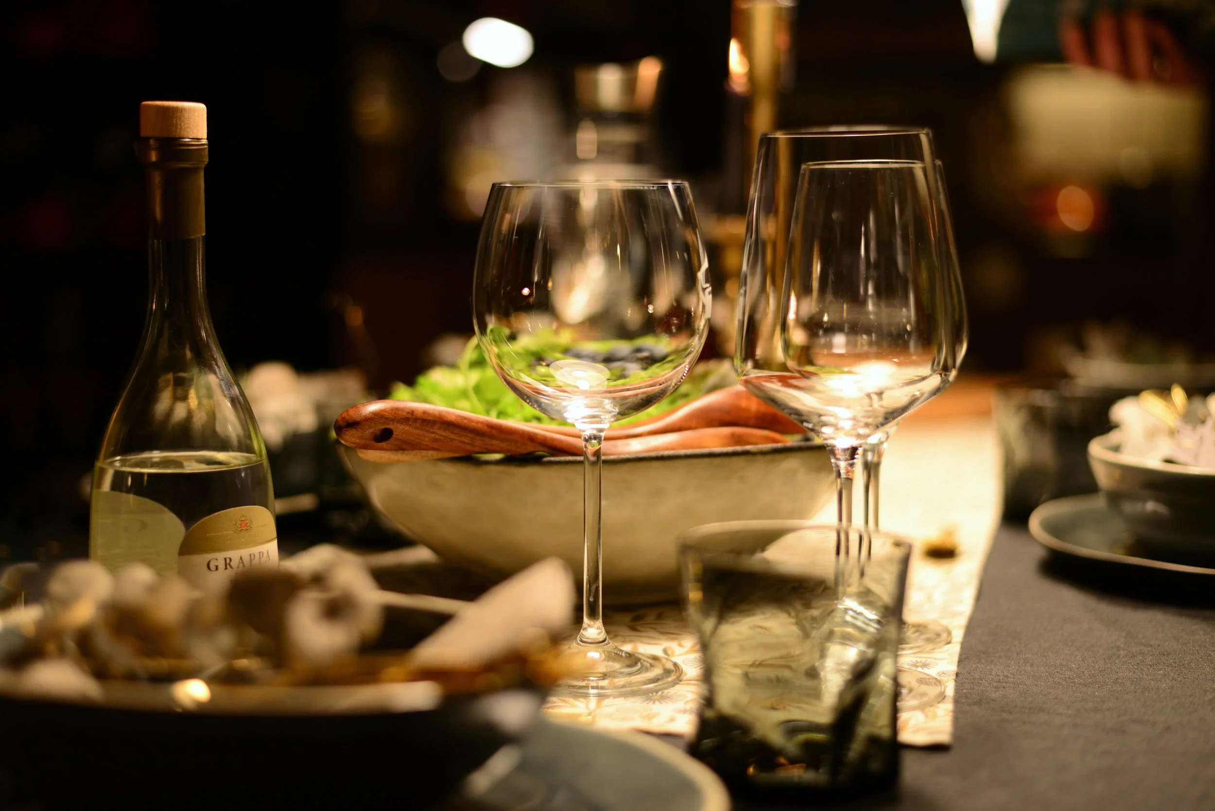 Close-up of empty wine glasses and a bottle of grappa on a dining table with a dark, warm ambiance.