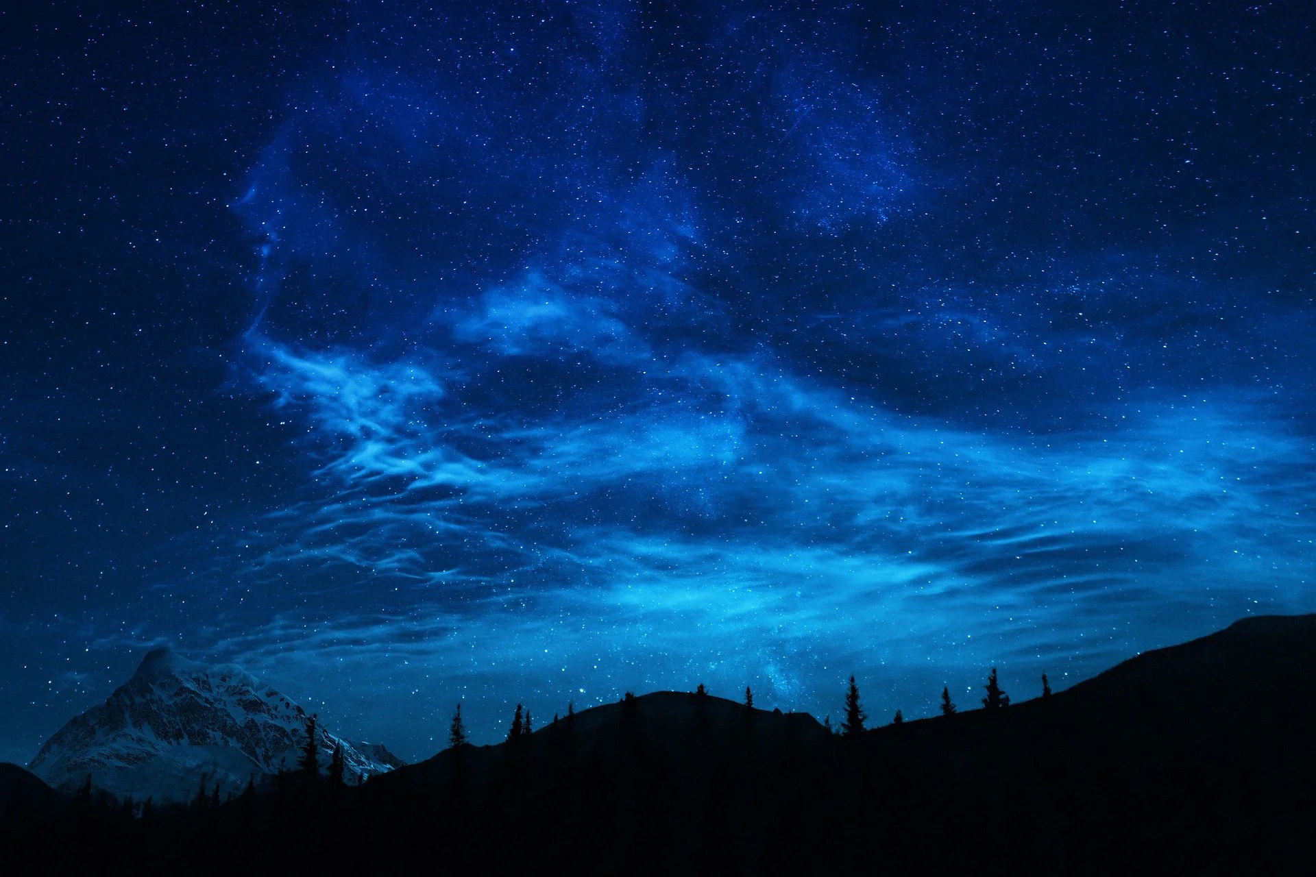 Night sky filled with stars and wispy clouds over a mountain range with snow-capped peaks and silhouetted trees.
