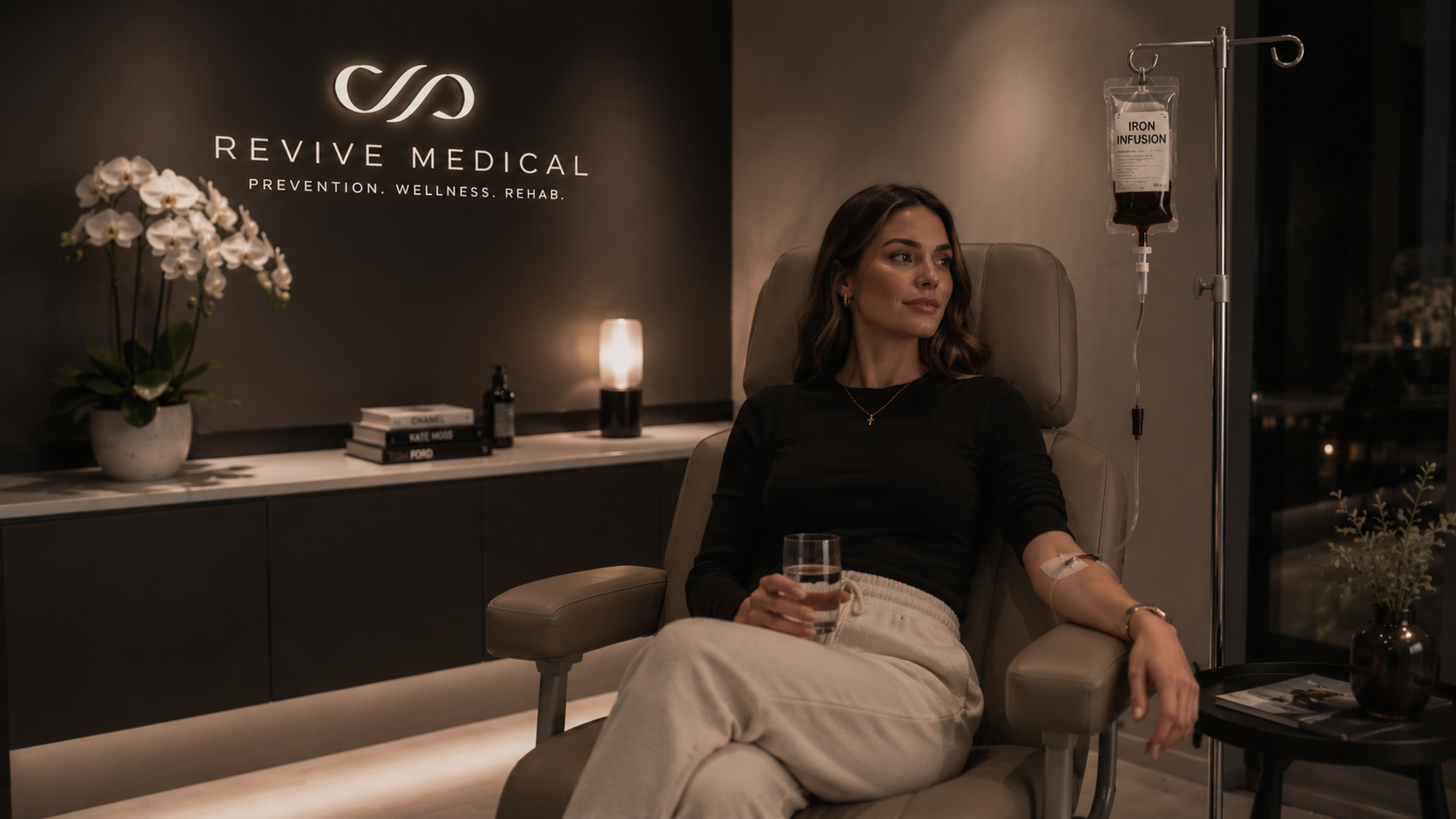 A woman receiving an iron infusion in a medical clinic with a modern interior. She is seated in a reclined chair, holding a glass of water, with a IV line connected to her arm. There is a stand with a bag of iron infusion fluid nearby.