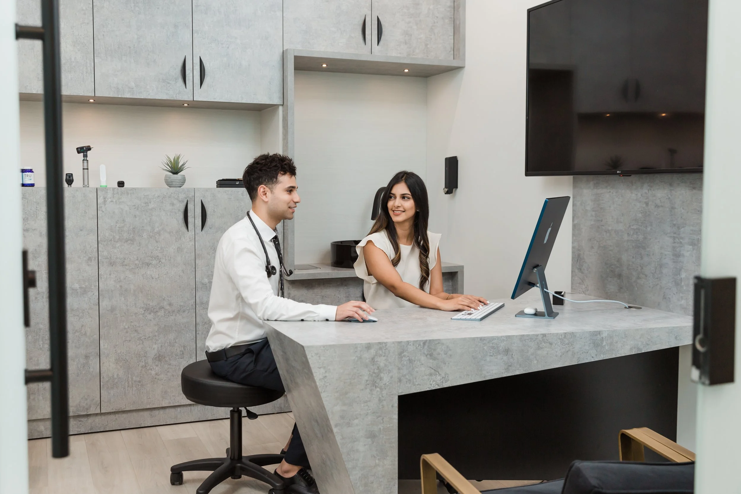 A doctor and a woman sitting at a reception desk in a modern medical office. The doctor has a stethoscope around his neck and is working on a computer, while the woman is smiling and looking at the doctor. The office has gray cabinets, a large TV, and a clean, minimalist design.